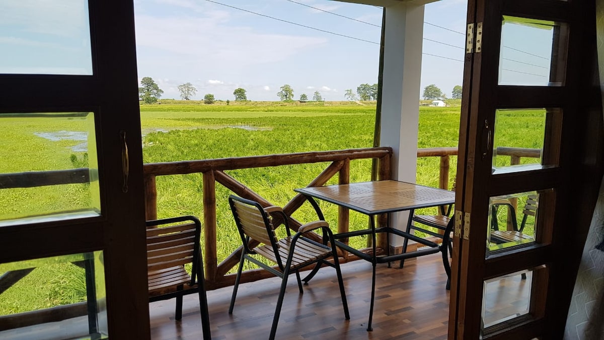 A balcony features a wooden railing and a table set with two chairs, providing a scenic view of the green paddy fields. The open space allows natural light to fill the area, while distant trees frame the landscape under a clear blue sky.