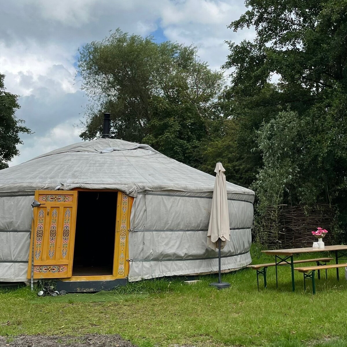 A cozy yurt is situated in a green outdoor setting, featuring a circular design and adorned with vibrant wooden doors. A patio umbrella stands beside a table with flowers, inviting relaxation in the serene surroundings. Lush trees create a peaceful backdrop.