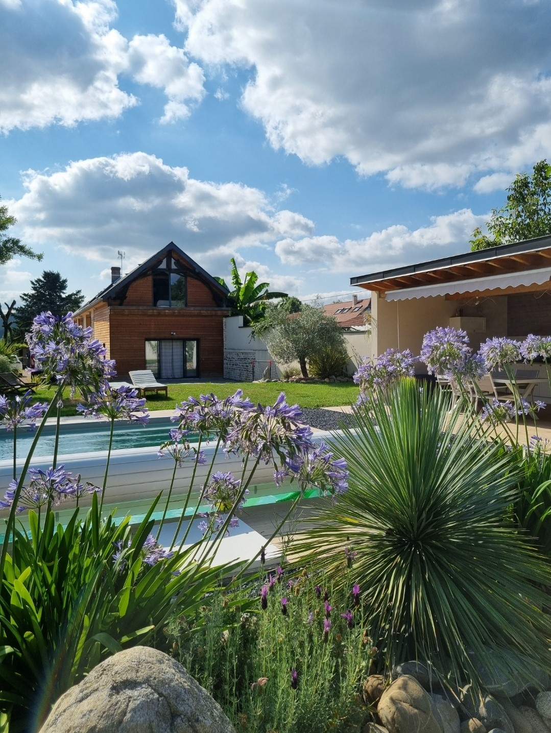 A well-maintained garden features vibrant purple flowers and lush greenery surrounding a swimming pool. A modern structure with a peaked roof is visible in the background, along with a partially shaded outdoor seating area. The sky is bright with scattered clouds.