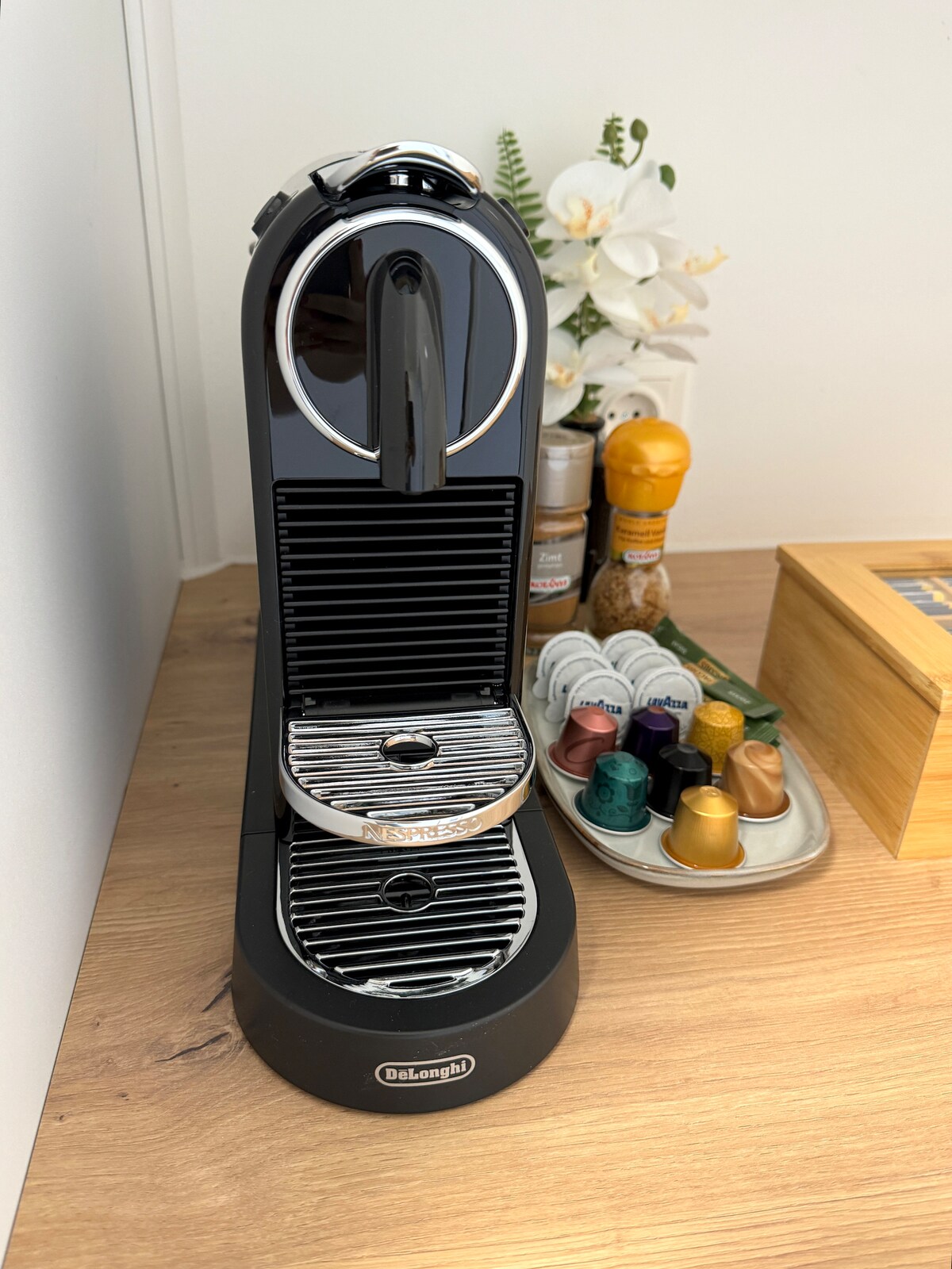 A modern Nespresso coffee machine is positioned on a light wooden countertop. Various coffee capsule colors are displayed on a tray nearby. A wooden box filled with utensils sits next to a small arrangement of white flowers, creating a functional and tidy coffee station.