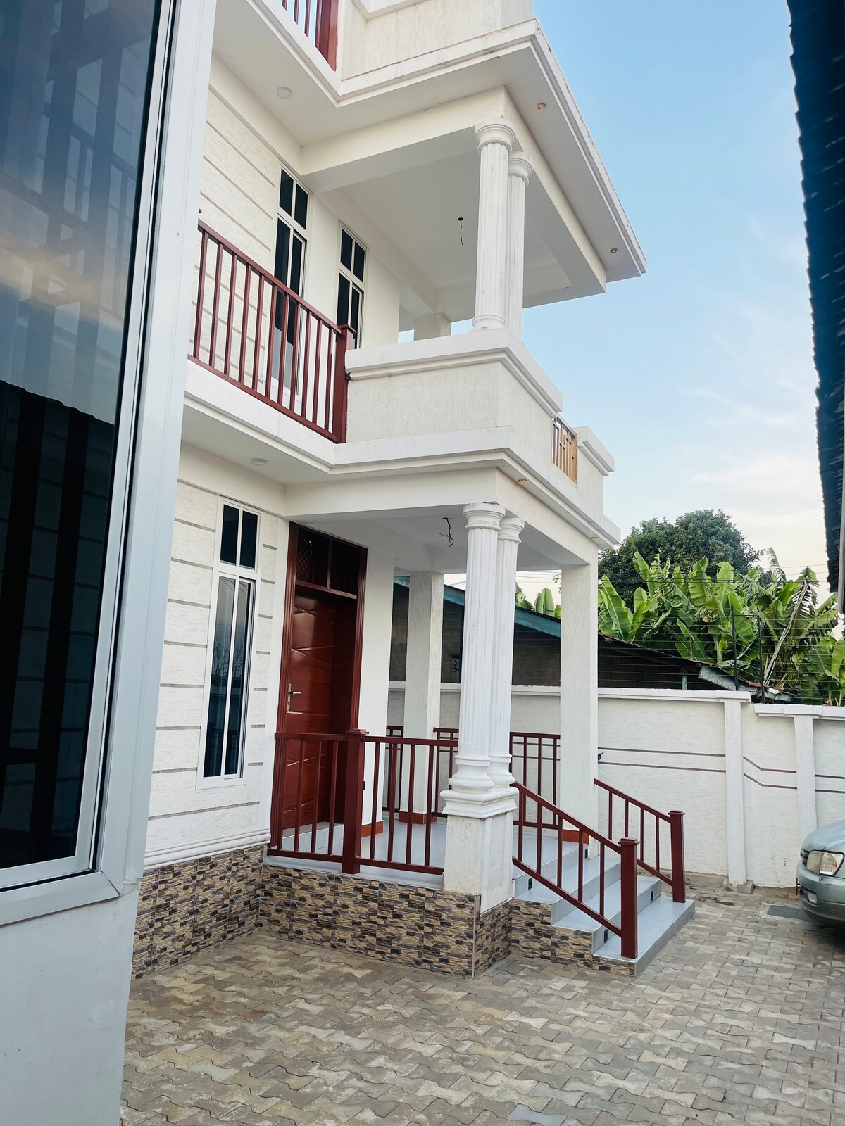An exterior view of the three-story apartment showcases a modern façade with a prominent entryway. A red railing encloses the staircase, leading to a decorative door. Paved stones are visible in the adjacent parking area, with lush greenery seen in the background.
