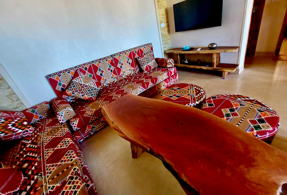 A modern living area is presented with a patterned chaise lounge and accent chairs, accompanied by a wide wooden coffee table. A flat-screen TV is mounted on the wall, while natural light brightens the space, enhancing its inviting feel.