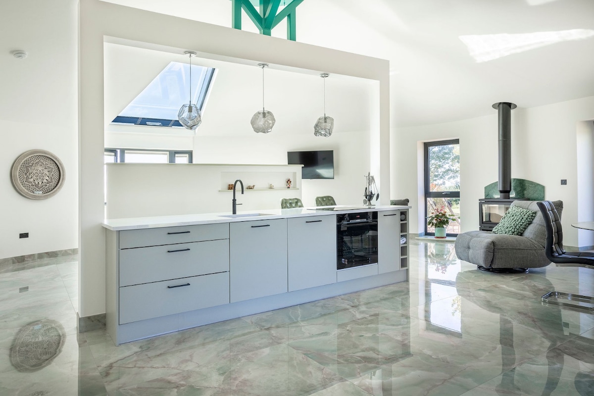 A modern kitchen design is showcased, featuring a sleek countertop with built-in appliances. Pendant lights illuminate the space, complemented by a skylight above. Reflective green marble flooring enhances the spacious feel. A comfortable seating area with a cozy chair is visible in the background.