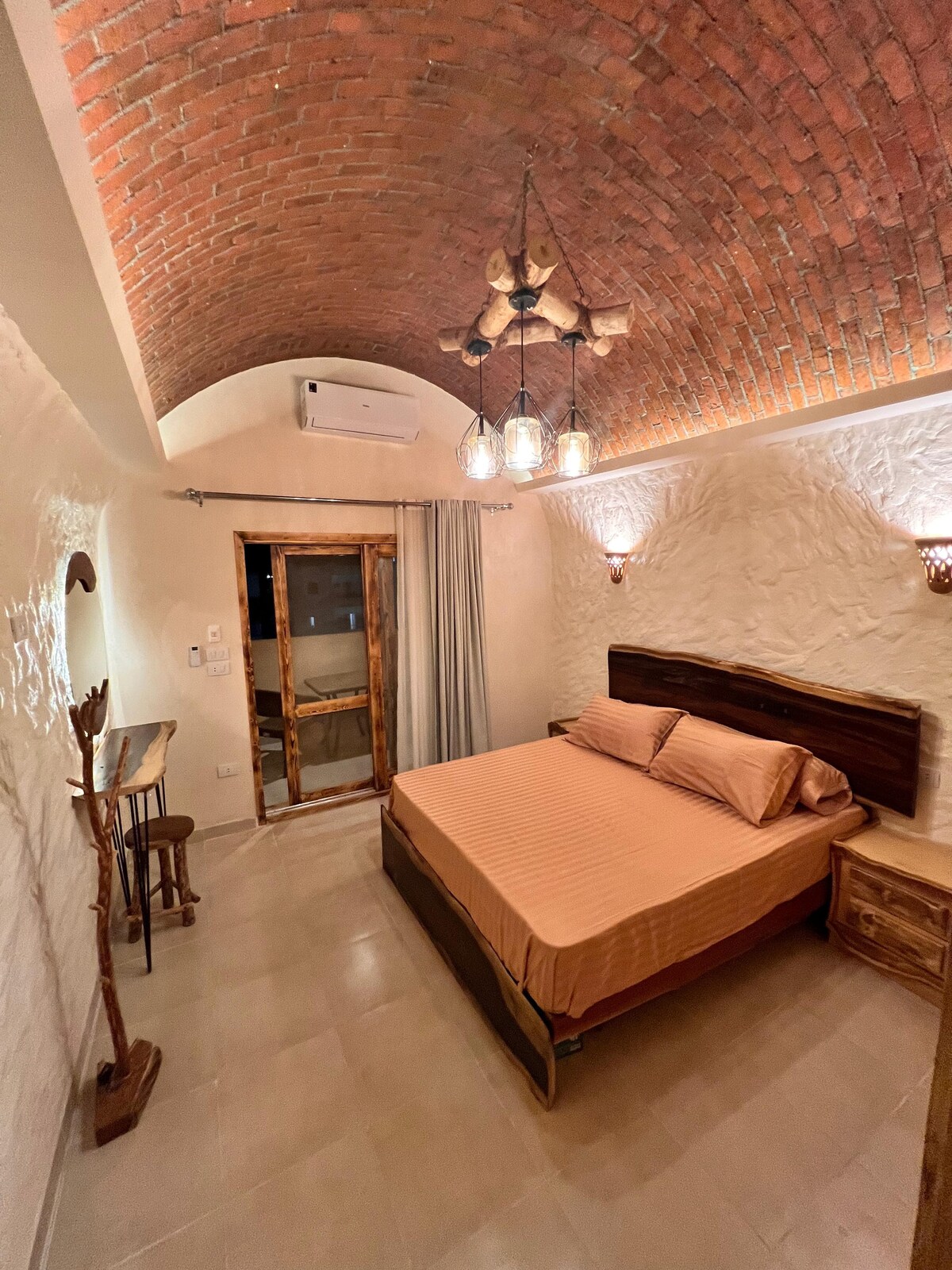 A cozy bedroom features a spacious bed with warm-toned bedding. Exposed brick arches accent the ceiling, and two pendant lights create a soft glow. A wooden door leads to a small terrace, while natural light enters through a nearby window, complemented by light curtains.