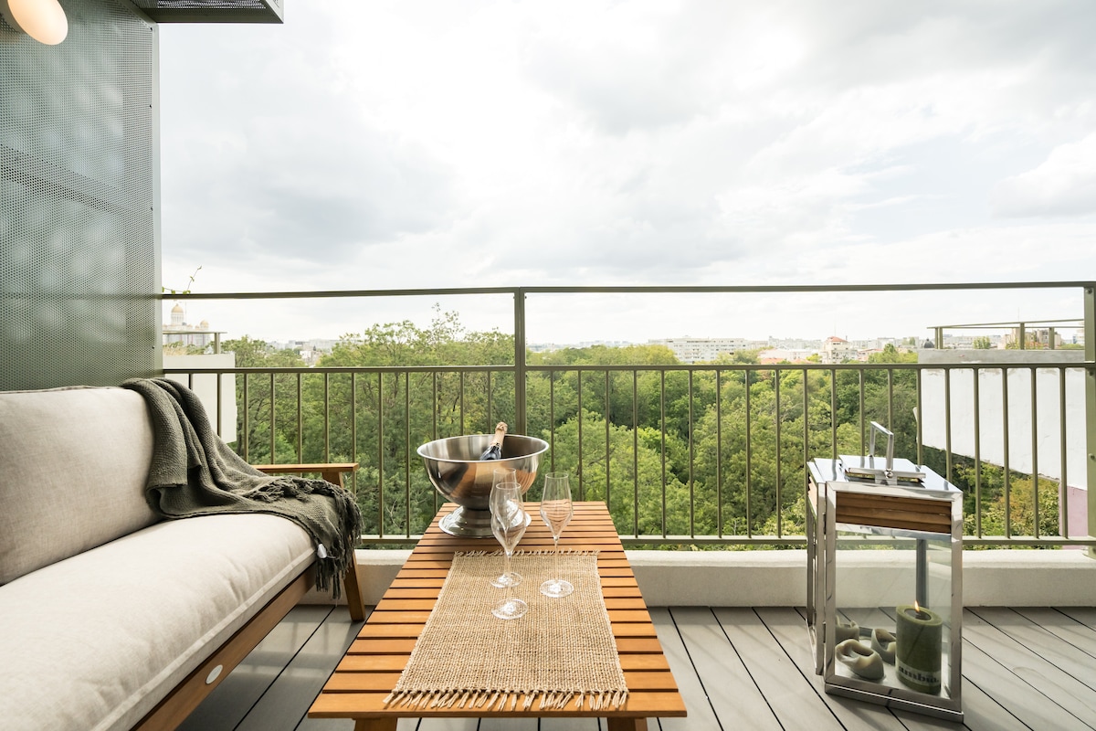 A spacious terrace features wooden furniture, including a lounge sofa and coffee table. A small serving cart holds decorative items. Lush greenery and city views can be seen in the background, creating a serene outdoor environment.