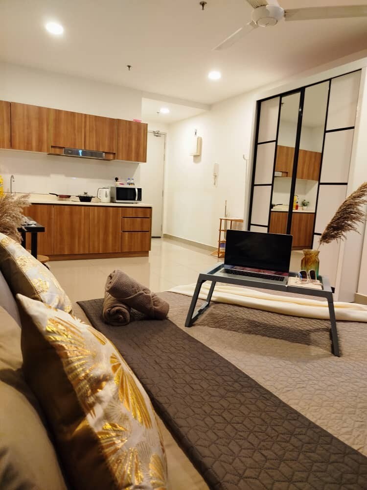 A modern living space is presented, featuring a neatly arranged couch with decorative pillows and a folded blanket. A coffee table sits in front, holding a laptop. The kitchenette is visible in the background, complemented by a mirror and light-colored walls that enhance the overall brightness.