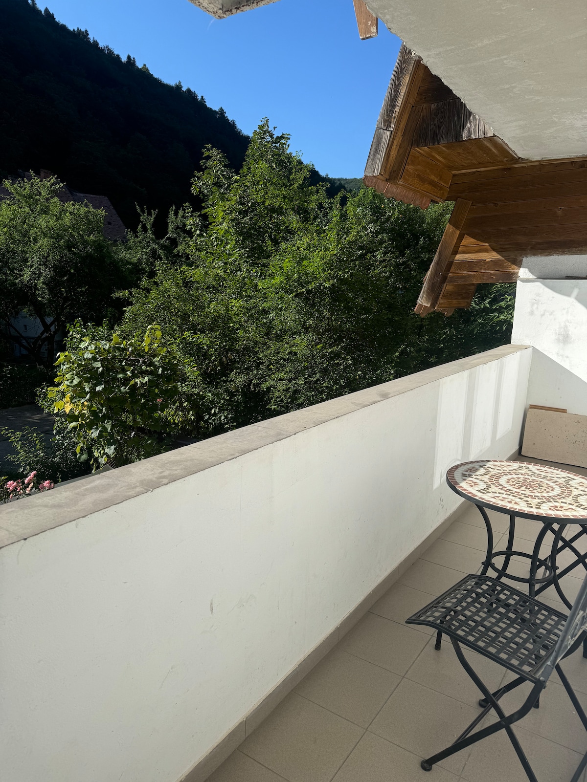 A small balcony is shown, featuring a round table with an intricate design and a pair of black metal chairs. Lush greenery from surrounding trees is visible, along with a clear blue sky, creating a serene outdoor space.