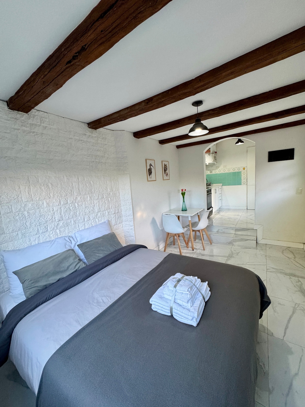 A cozy sleeping area features a spacious bed with neutral bedding, complemented by a woven throw. Wooden beams adorn the ceiling, and natural light brightens the room. A small dining table with two chairs is visible nearby, enhancing the inviting layout.