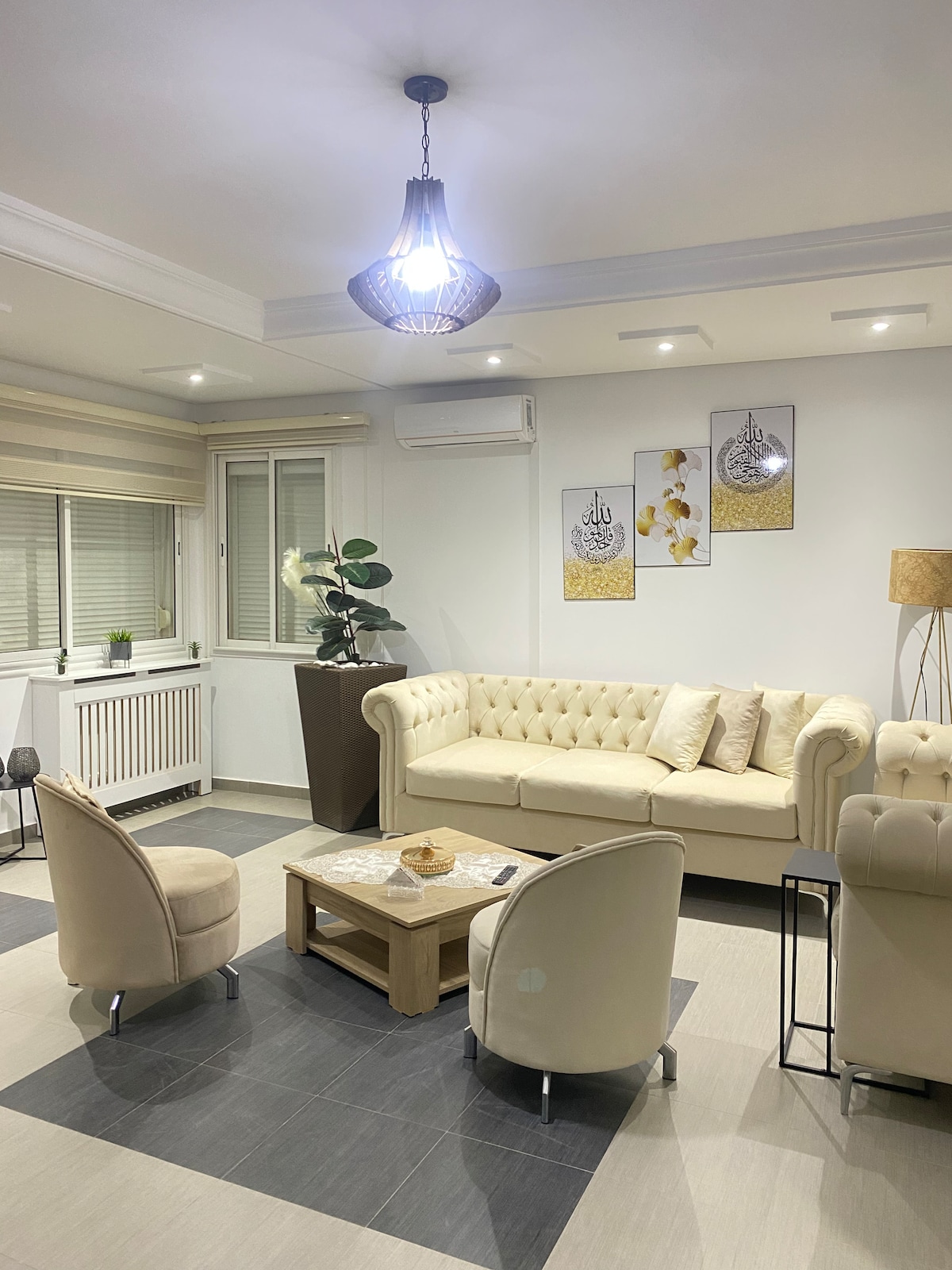 A cozy living area is furnished with a light-colored tufted sofa, accompanied by two armchairs and a wooden coffee table. Artwork adorns the wall, and a potted plant adds a touch of greenery. Natural light enters through large windows, complemented by soft ambient lighting.