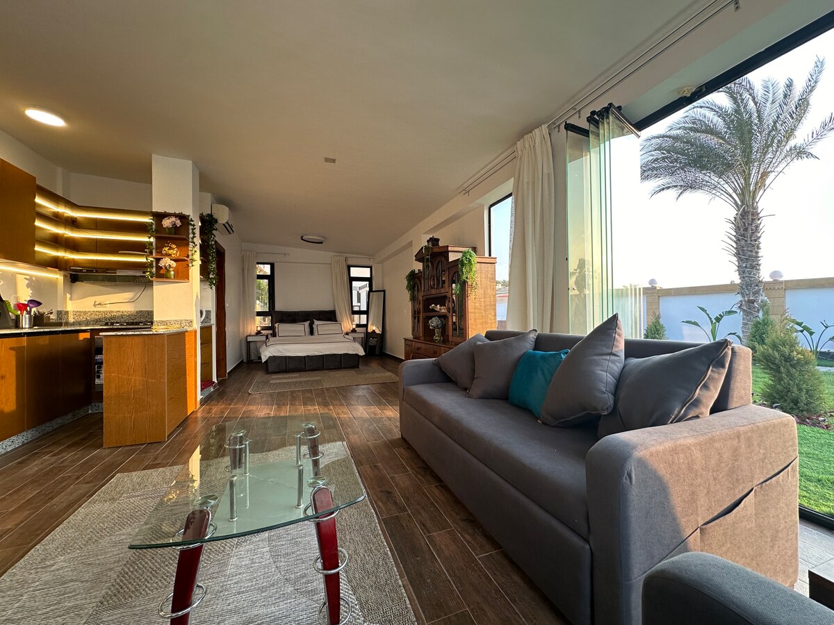 An open living area features modern furnishings, including a spacious gray sofa and a glass coffee table, while natural light floods in through large windows. In the background, a comfortable sleeping area is visible, complemented by soft bedding and a view of the garden.