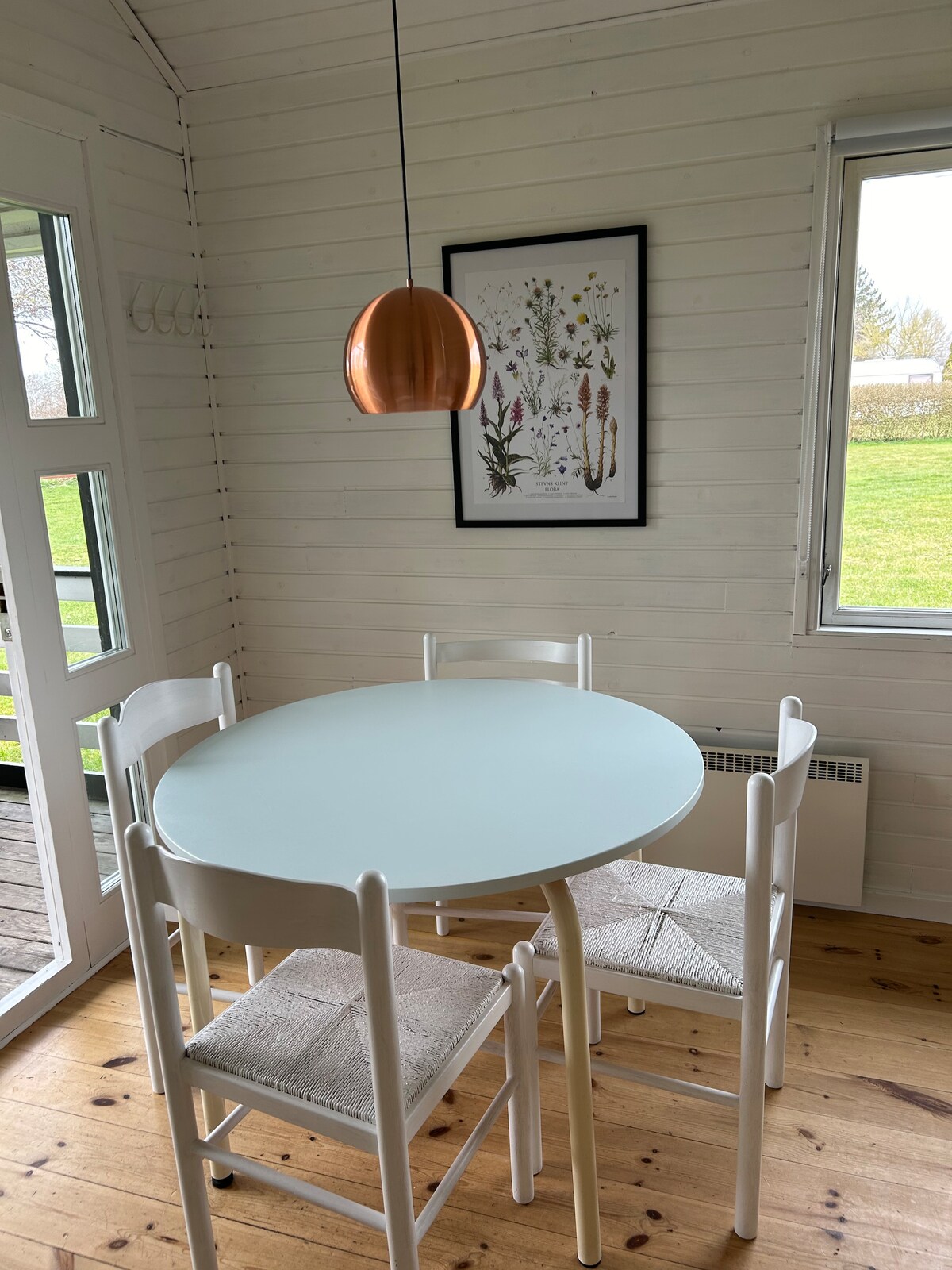 A round dining table with a light blue surface is accompanied by four white chairs, each featuring a woven seat. A copper pendant light hangs above the table, creating a simple yet inviting dining space with large windows that let in natural light.