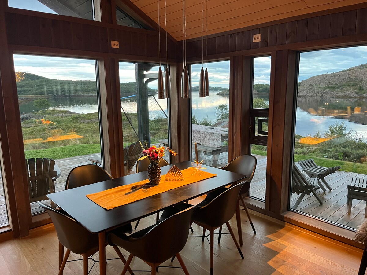 A dining area features a sleek black table surrounded by stylish chairs, illuminated by pendant lighting. Large glass windows provide views of the serene water landscape outside, while a wooden deck and seating area can be seen beyond. A colorful arrangement of flowers sits on the table.