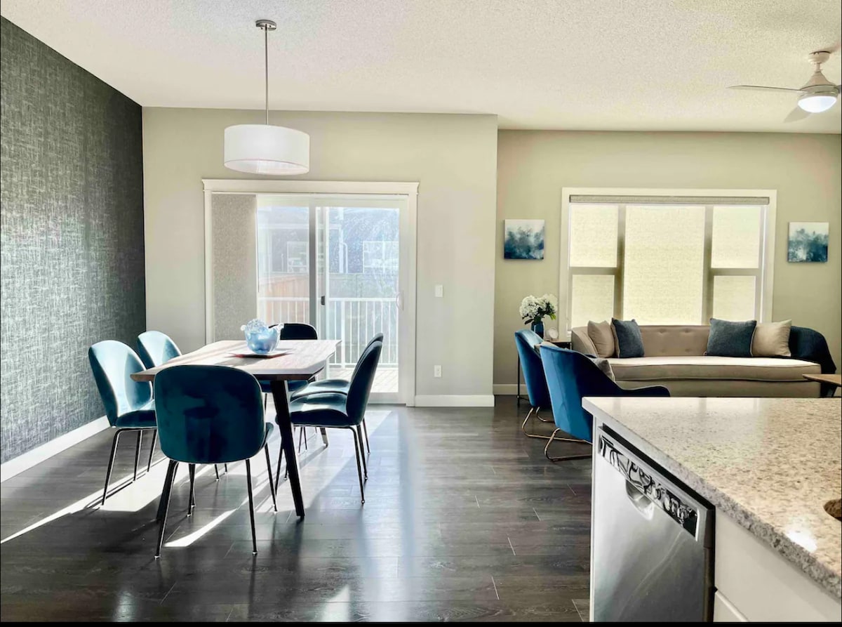 A dining area is visible, featuring a medium-sized table surrounded by six upholstered chairs. A contemporary light fixture hangs above the table. Natural light floods the space, highlighting the nearby seating area with a sofa and decorative cushions. Large windows provide views of the outside.