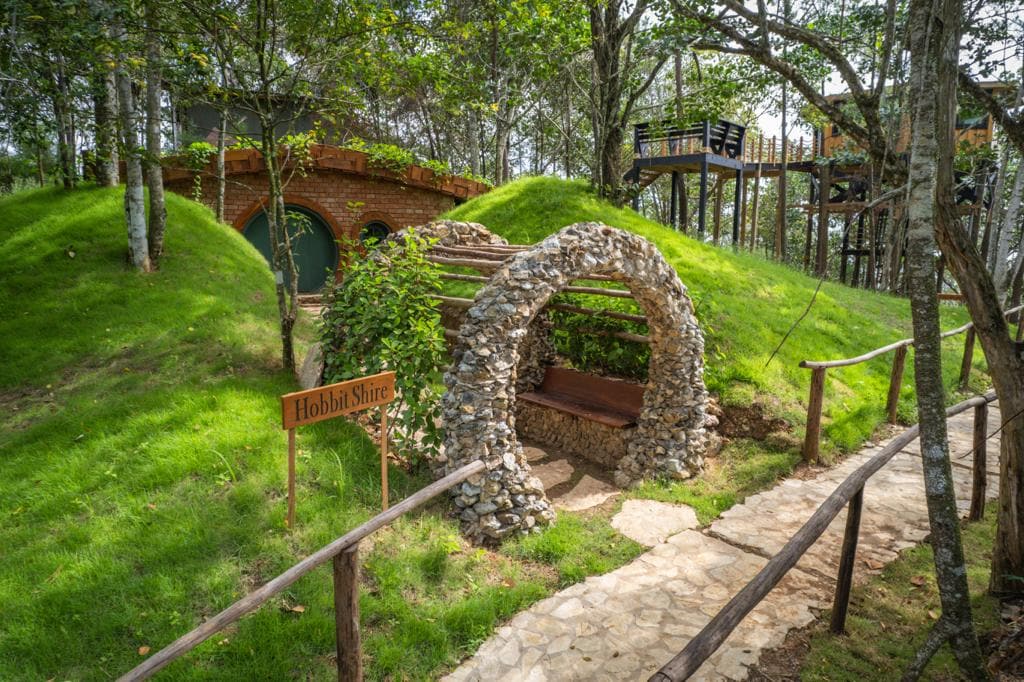 A stone archway leads to a cozy resting area amid lush greenery. The pathway is bordered by neatly trimmed grass, creating a natural setting. A wooden sign reads 'Hobbit Shire,' indicating a whimsical touch to the surroundings.