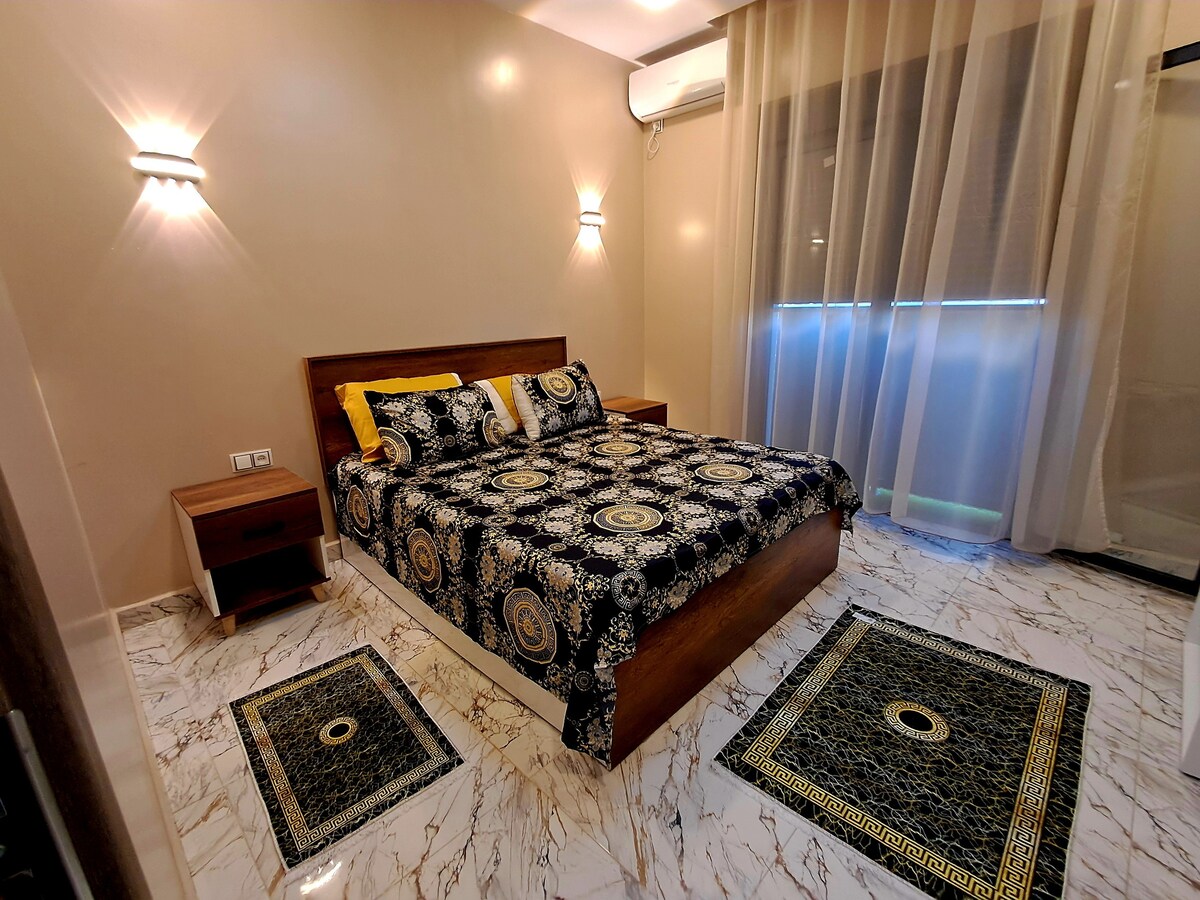 A spacious sleeping area features a patterned bedspread on a wooden platform bed. Soft lighting is provided by wall sconces, and sheer curtains allow natural light to filter through. Decorative rugs complement the marble floor, creating a cohesive design.