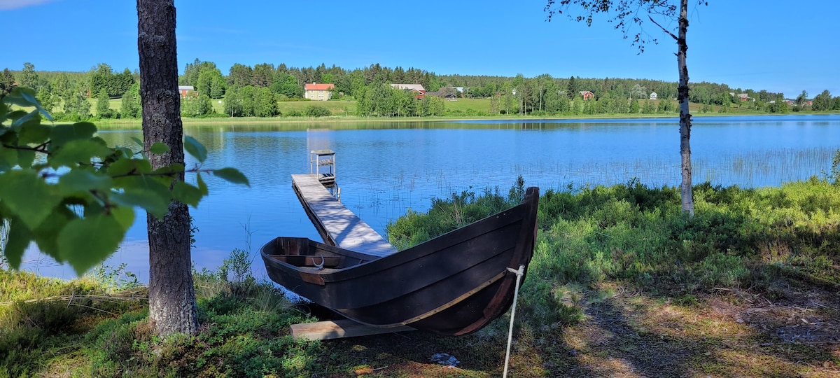 A tranquil lakeside scene features a wooden rowboat resting near a private dock. Lush greenery frames the water, while gently rolling hills extend into the background, creating a serene environment. The clear sky reflects off the smooth lake surface.