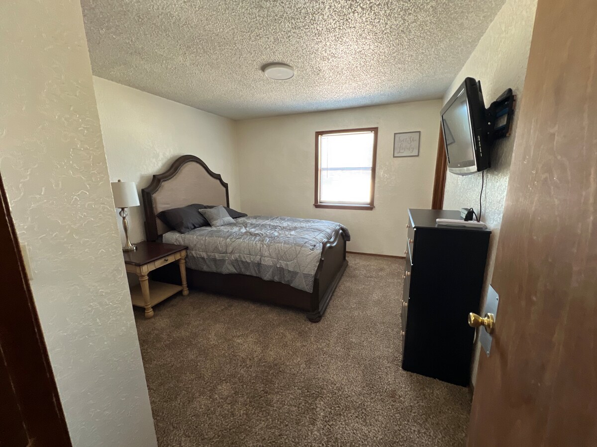 A comfortable bedroom is presented, featuring a spacious bed covered with a neatly arranged quilt. A bedside table with a lamp is positioned next to the bed. A dresser is situated across from the bed, and a flatscreen TV is mounted on the wall. Natural light enters through the window.