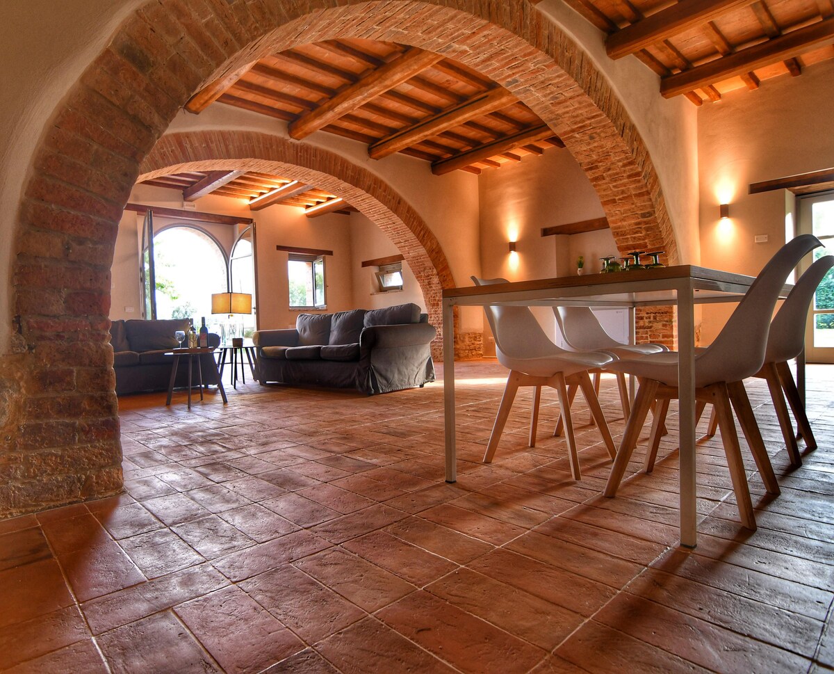An expansive vaulted living room is featured, showcasing terracotta flooring and exposed wooden beams. Natural light filters through arched windows, illuminating the seating area with plush sofas and a light-colored dining table. The inviting space highlights an atmosphere conducive to relaxation.