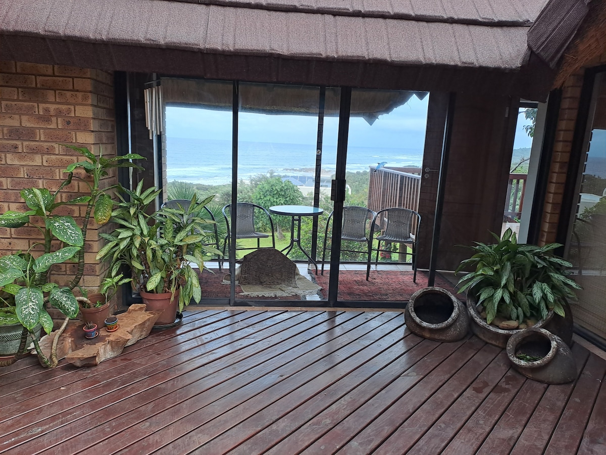 Large glass doors open to reveal a spacious balcony overlooking the ocean. Potted plants and decorative arrangements are placed on a wooden floor, creating a connection between indoor and outdoor spaces while highlighting the coastal view.