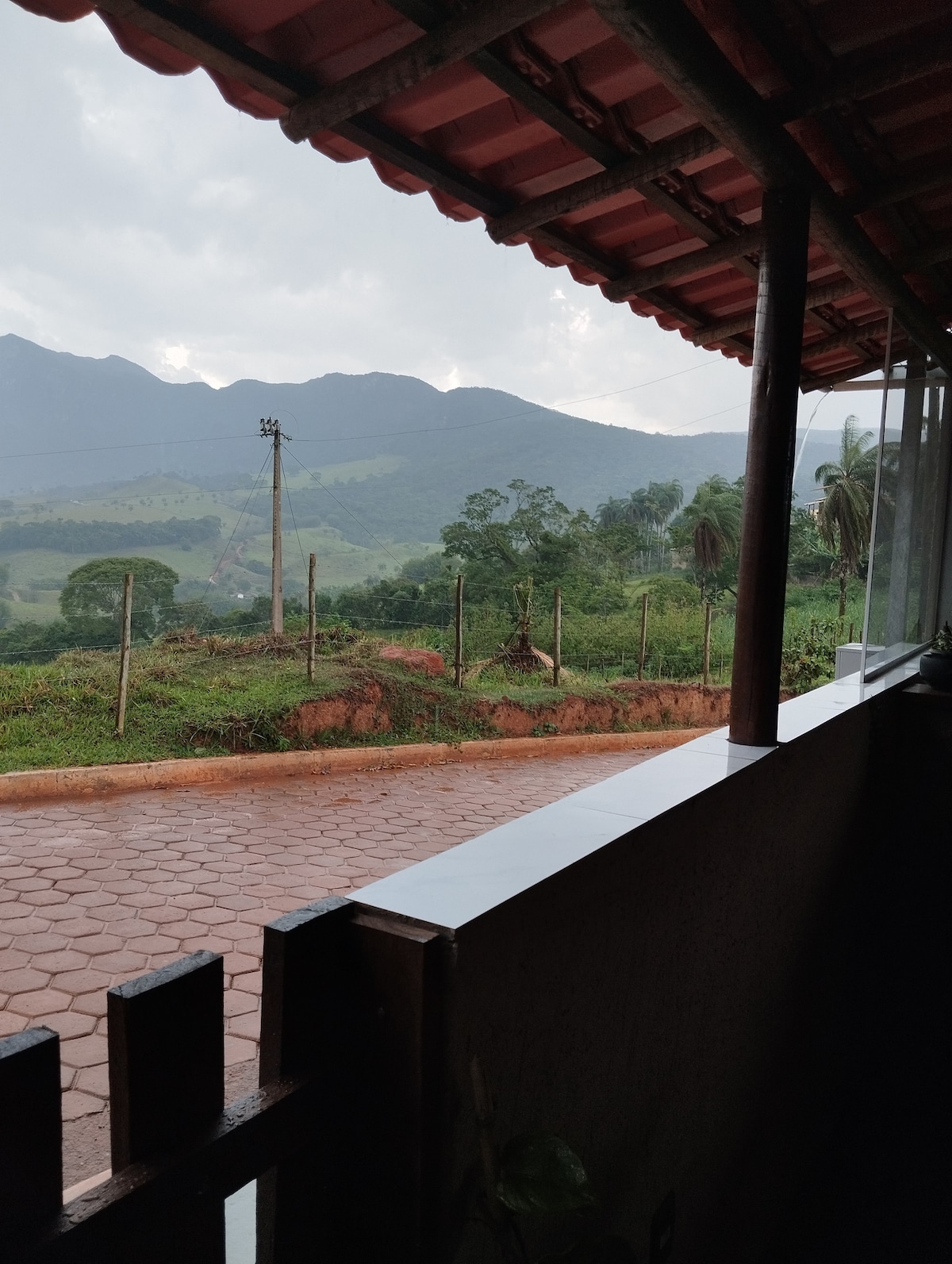A covered outdoor area features a solid railing, overlooking a lush green landscape. Rolling hills and distant mountains are visible under a cloudy sky. The terrain shows a mix of trees and open spaces, creating a serene environment.