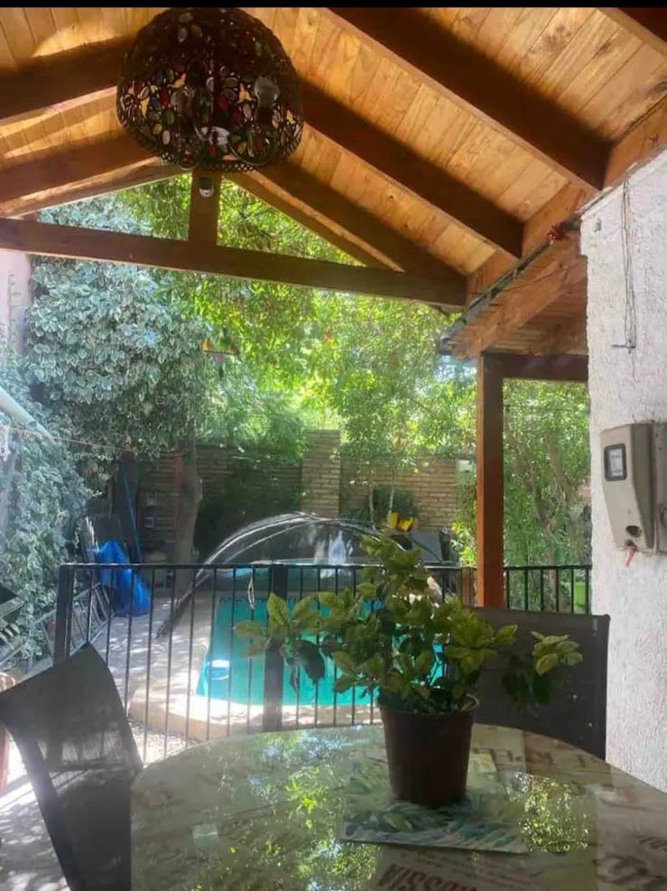 A shaded outdoor dining area features a round table with a potted plant at its center. Lush greenery surrounds the space, providing a natural backdrop. A swimming pool is visible through the gated entry, framed by trees and the warm wood of the ceiling.
