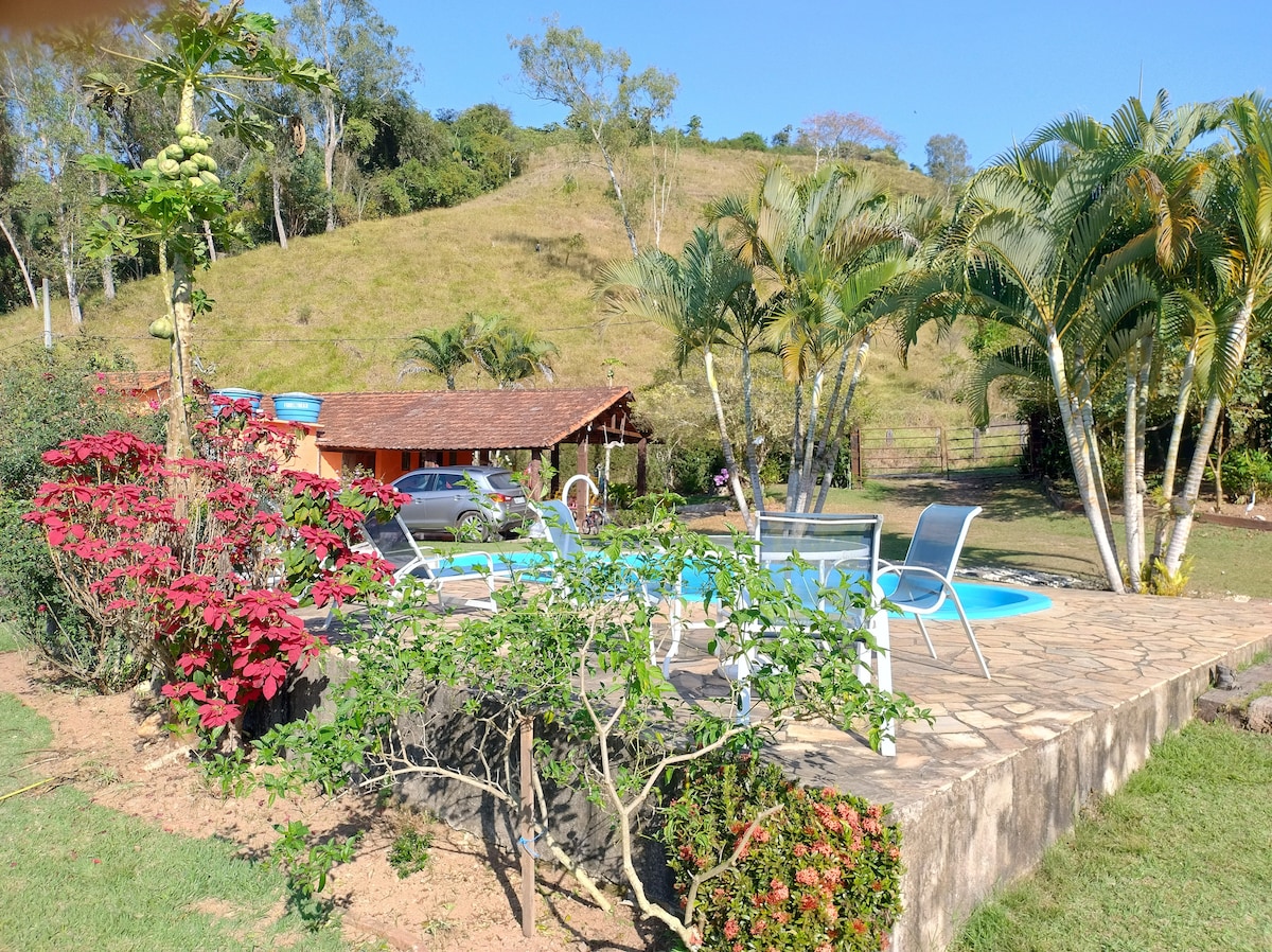 A serene outdoor space features a refreshing pool surrounded by lush greenery and flowering plants. Comfortable lounge chairs are positioned on a stone patio, offering a perfect spot for relaxation. In the background, a charming rustic building is nestled against a hillside, enhancing the natural scenery.