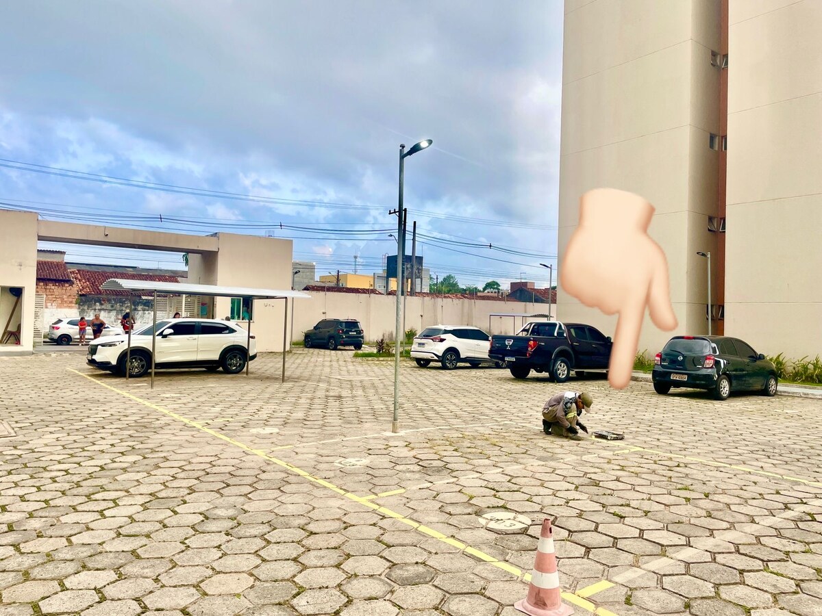 A spacious parking lot is visible, featuring several vehicles parked in designated spaces. A person is seen maintaining the area, with modern buildings in the background and a cloudy sky overhead. Streetlights provide illumination, enhancing the area’s accessibility.