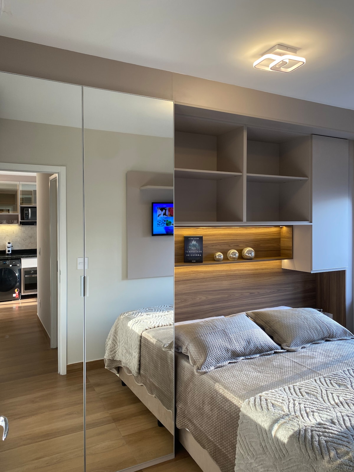 A modern bedroom features a spacious bed dressed in soft linens. A mirrored wardrobe reflects the room's dimensions. Above the bed, a wooden shelf displays decorative items. Soft lighting highlights the neutral tones, creating a serene environment. The entrance to a kitchen area is visible in the background.