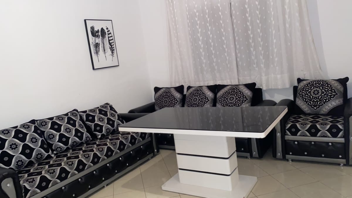 A cozy sitting area features a dark patterned sofa and a contrasting armchair, both adorned with decorative cushions. A sleek, modern dining table sits at the center, complemented by a light fabric curtain that gently diffuses natural light from the window.