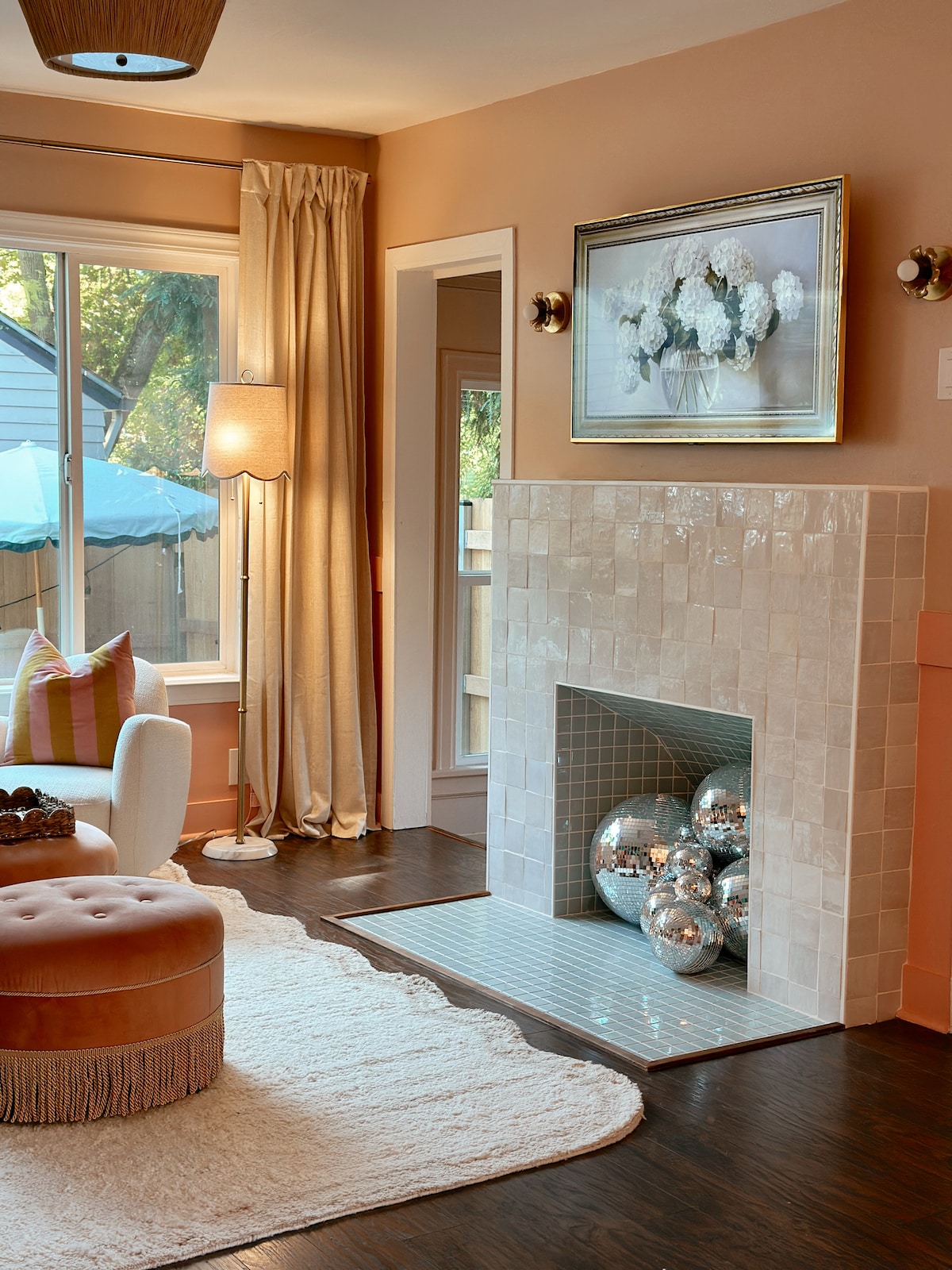 A cozy living room features a light-colored area rug and a plush, circular ottoman with fringe. A decorative fireplace with shimmering silver spheres is set against a tiled backdrop. Natural light streams through a large window adorned with flowing curtains, illuminating the inviting space.