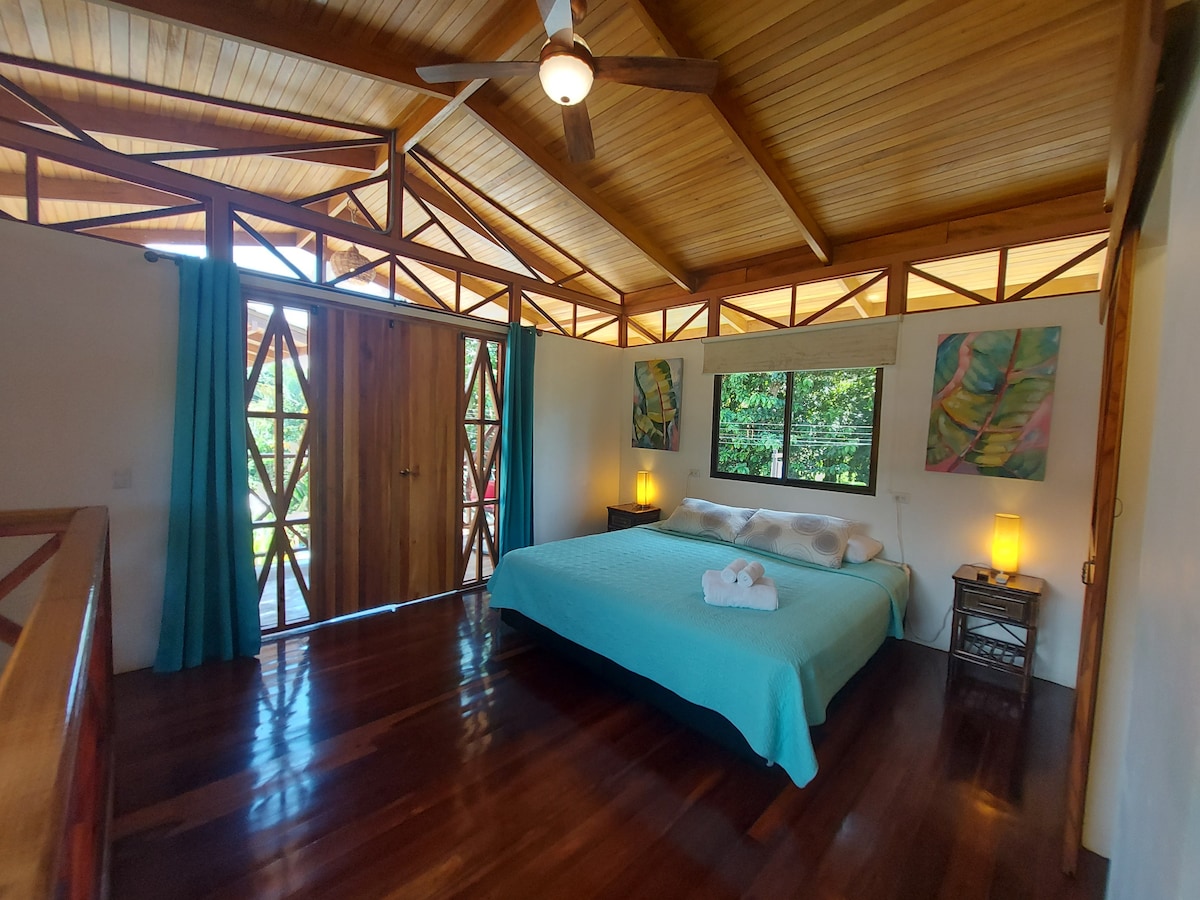 A spacious bedroom is equipped with a king-size bed and features wooden floors. Two bedside tables with lamps add soft lighting. Large windows provide views of the surrounding greenery. Colorful artwork decorates the walls, and soft turquoise curtains frame the windows, enhancing the natural light.