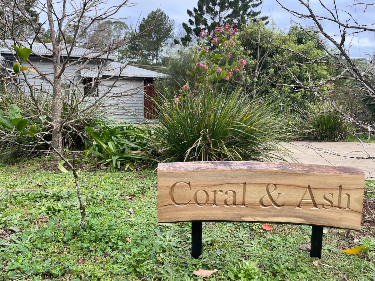 A wooden sign displaying 'Coral & Ash' stands prominently in the foreground, surrounded by lush greenery and flowering plants. The exterior of the property is partially visible in the background, showcasing a simple architectural style that blends with the natural surroundings.