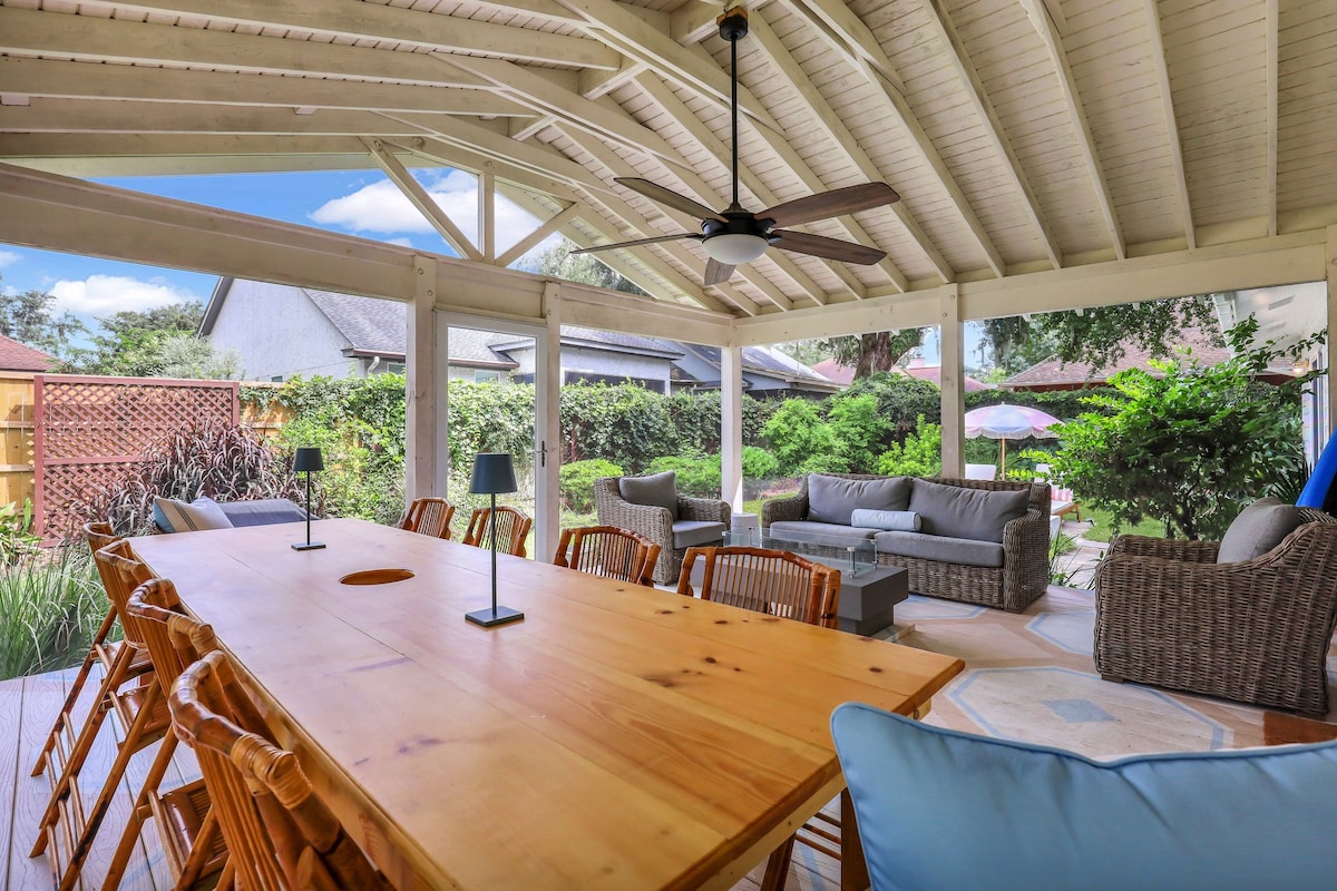 A spacious screen porch features a large wooden dining table surrounded by eight chairs, ideal for gatherings. Comfortable couches offer plush seating options, and a ceiling fan hangs above, providing a cool ambiance. Lush greenery surrounds the outdoor area, enhancing the serene environment.