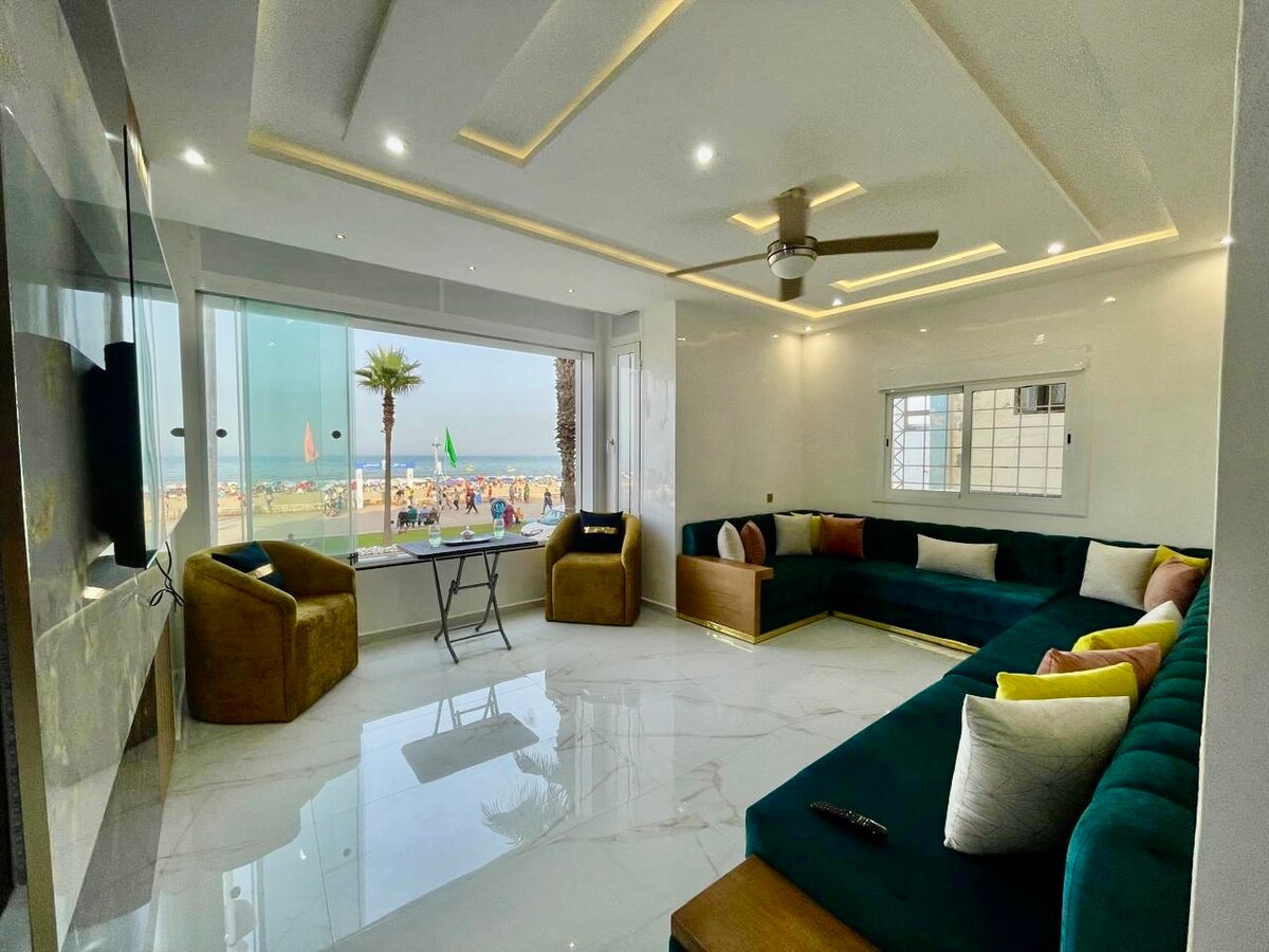 A spacious living area features large glass windows that offer views of the beach. The room contains a comfortable sectional sofa with an array of colorful cushions. A sleek coffee table is positioned in the center, while a ceiling fan provides ventilation.