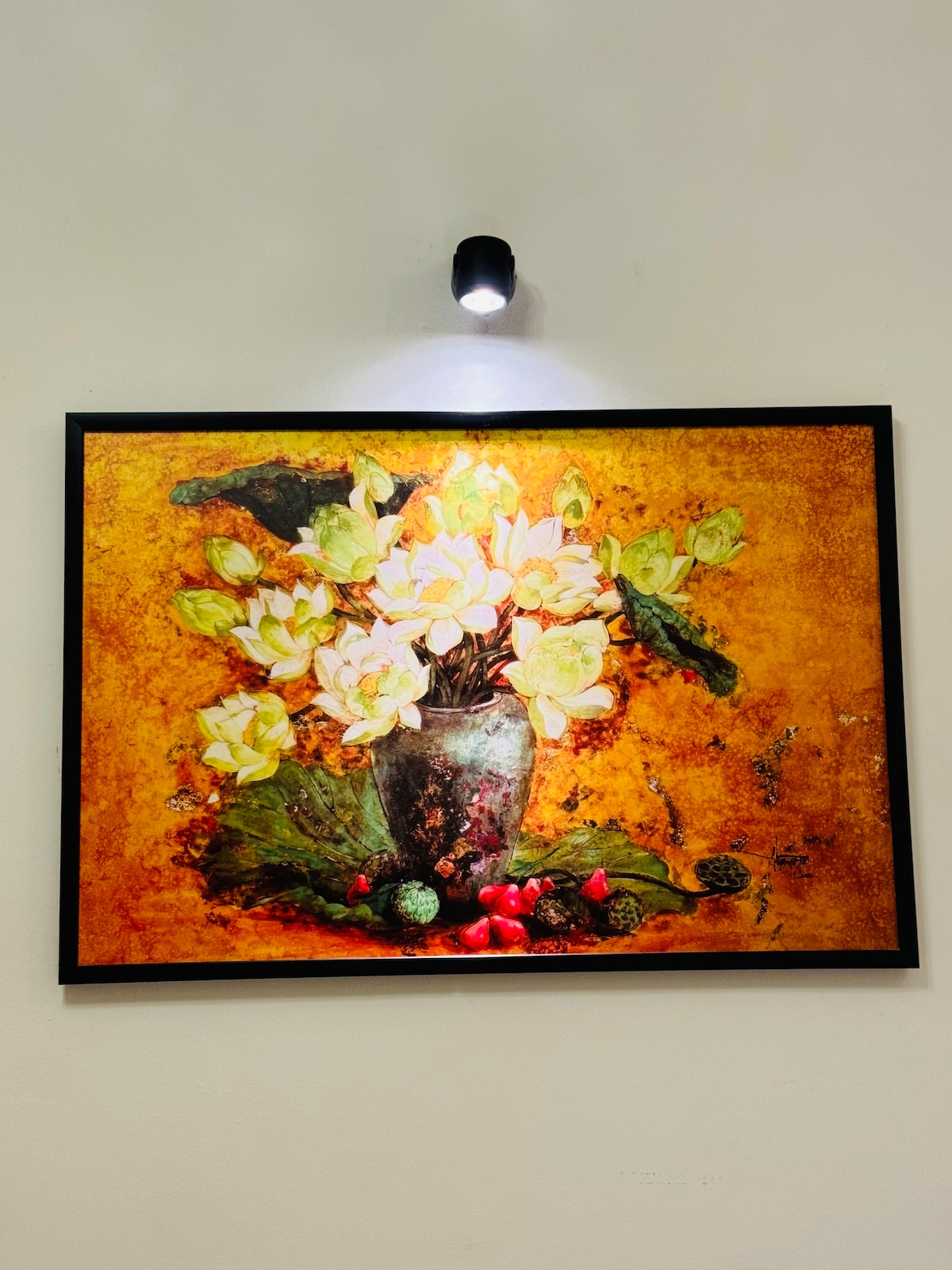 A framed artwork of a floral arrangement is displayed on a wall. The painting features white flowers in a dark vase, set against an orange background. Subtle lighting highlights the intricate details, creating a focal point in the space.
