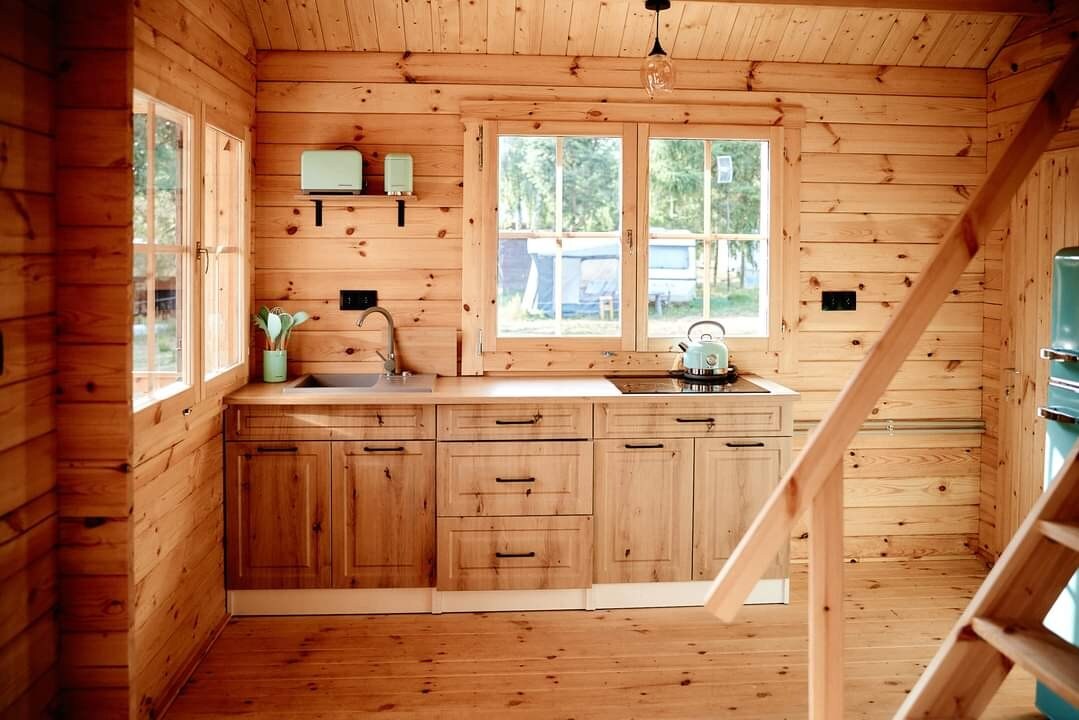 A wooden kitchen space is featured within the cabin, showcasing natural wood cabinetry and a stainless steel sink. Light floods in through the large windows, complementing the wooden interior and providing a view of the outdoor surroundings. An electric kettle and modern cooking appliances are visible.