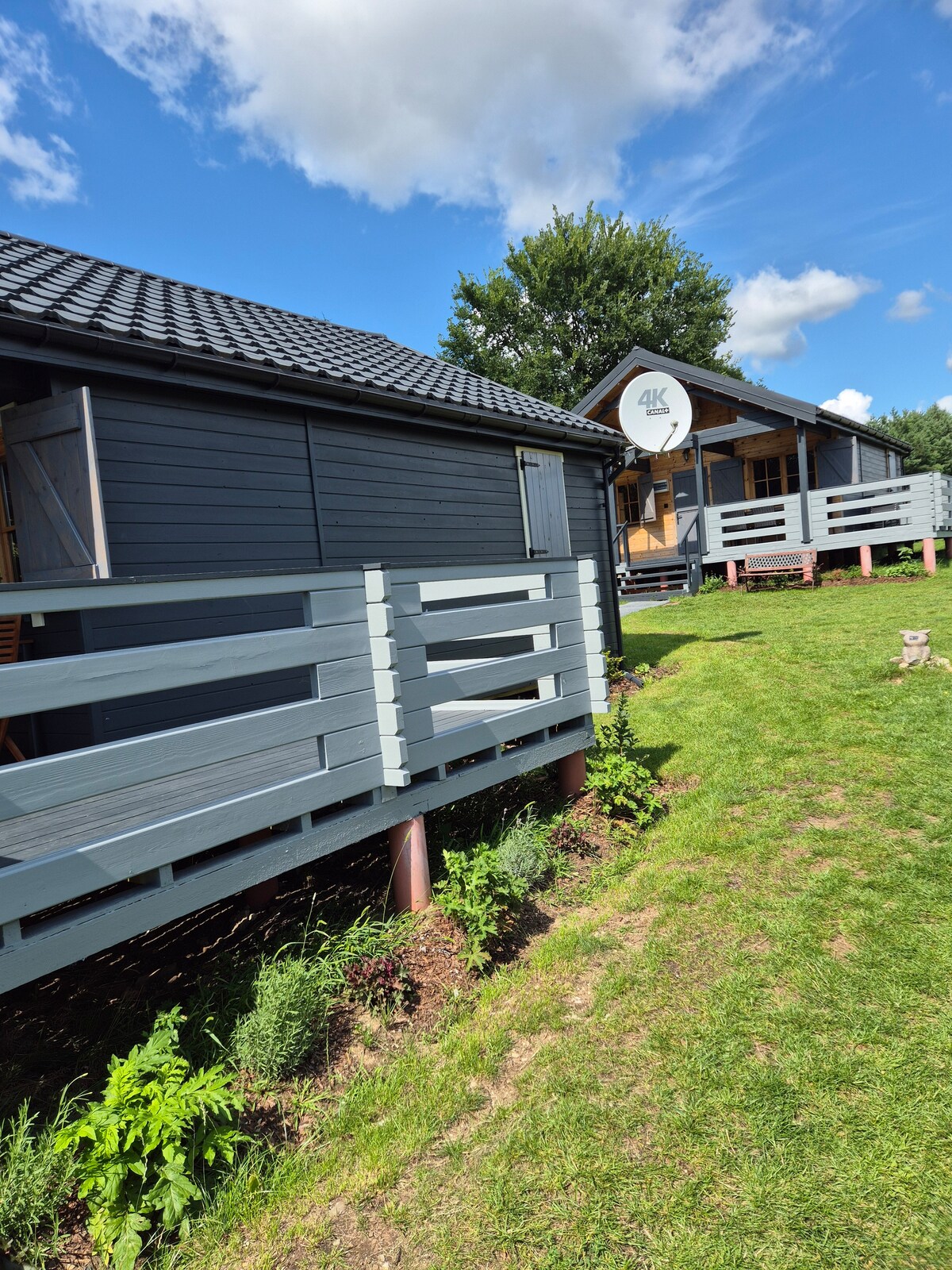 Two wooden cabins are visible against a clear blue sky, surrounded by lush green grass. Each cabin features a gray exterior with a covered porch. Simple wooden railings are present, creating a welcoming outdoor space. A small decorative feature can be seen in the foreground.