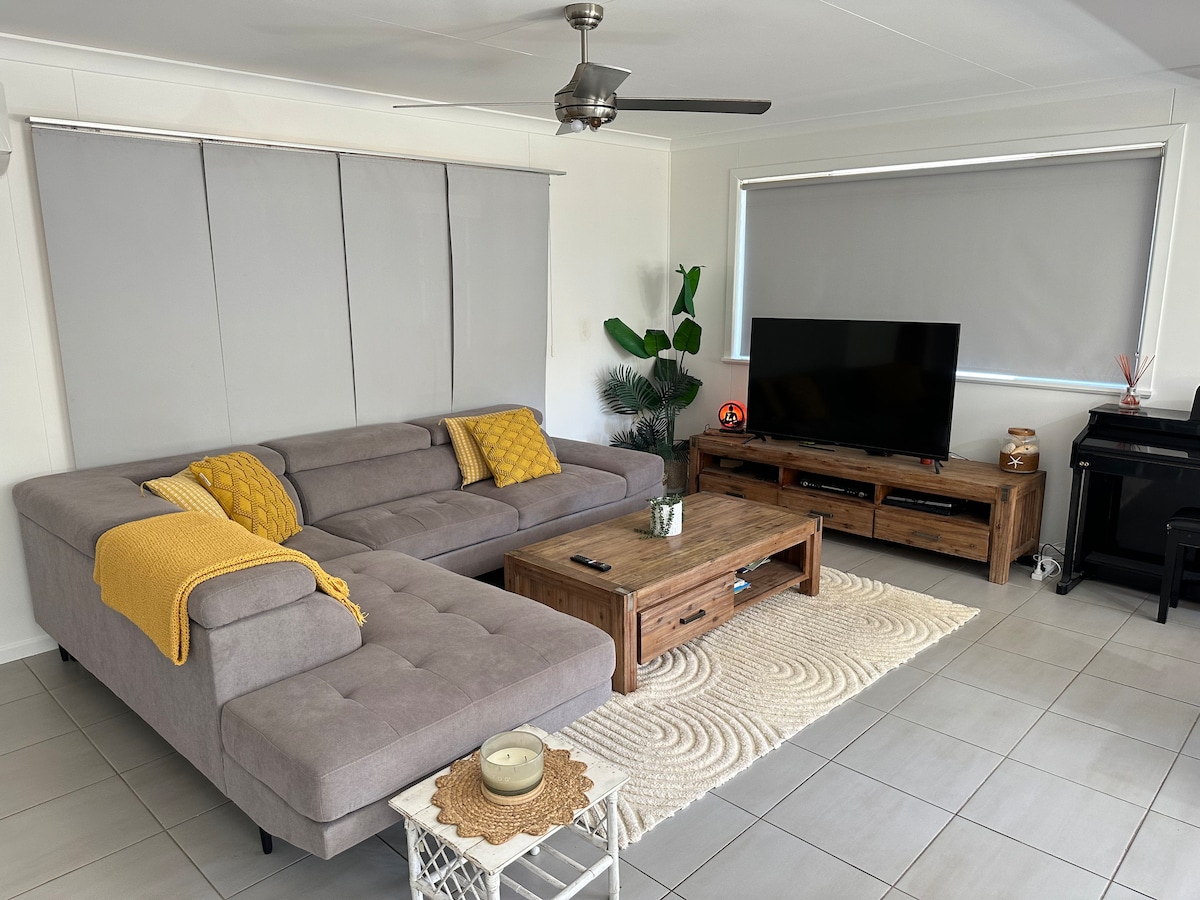A modern living room is showcased with a spacious gray sectional sofa, embellished with a yellow throw blanket. A wooden coffee table sits in the center, accompanied by a textured rug. A flat-screen TV is positioned on a wooden entertainment unit, with natural light filtering through the window.