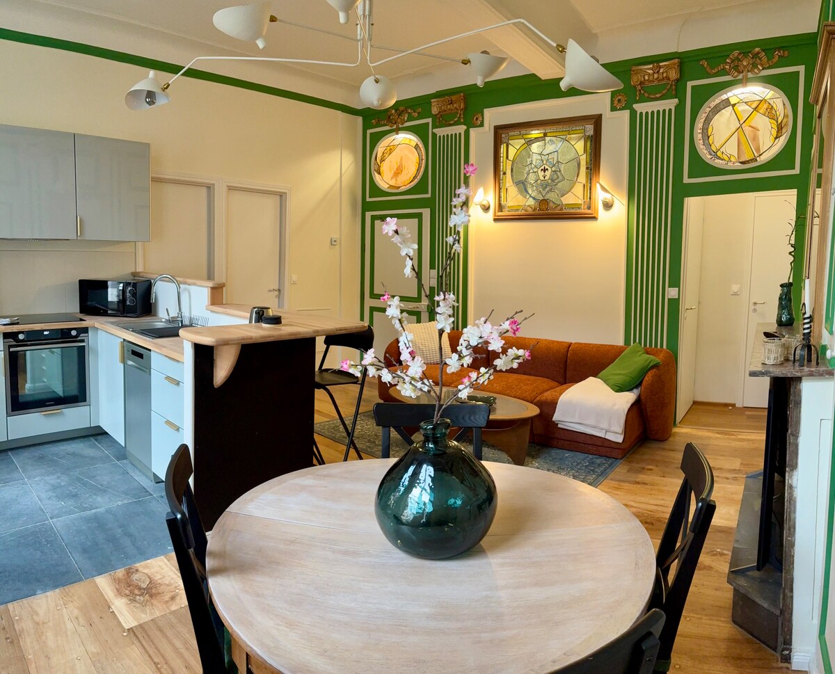 A spacious living area is captured, featuring an inviting brown sofa and a round dining table surrounded by dark chairs. A modern kitchen with sleek cabinetry is visible, alongside decorative wall details and large circular artworks. A vase with branches adds a natural touch to the dining table.