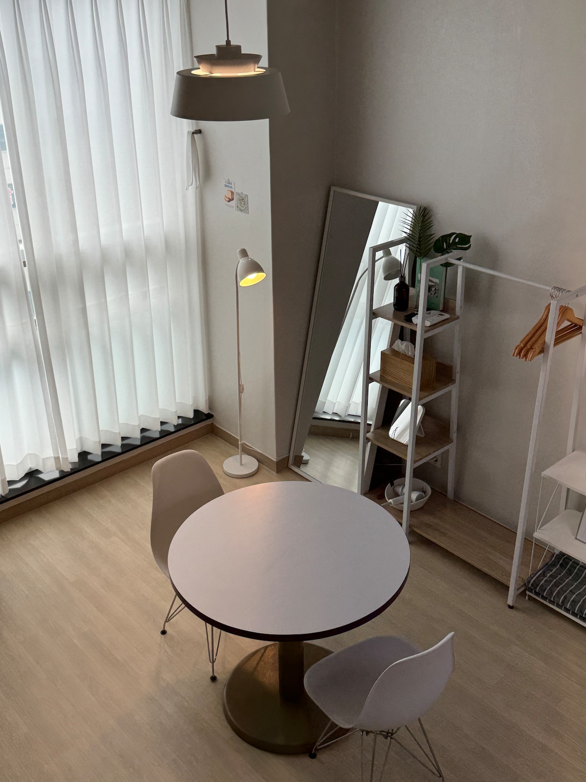 A dining area is visible, featuring a round table accompanied by two chairs. A tall mirror reflects the light-filled space, while a standing lamp provides additional illumination. A shelving unit displays decorative items, and sheer curtains allow natural light to filter in.