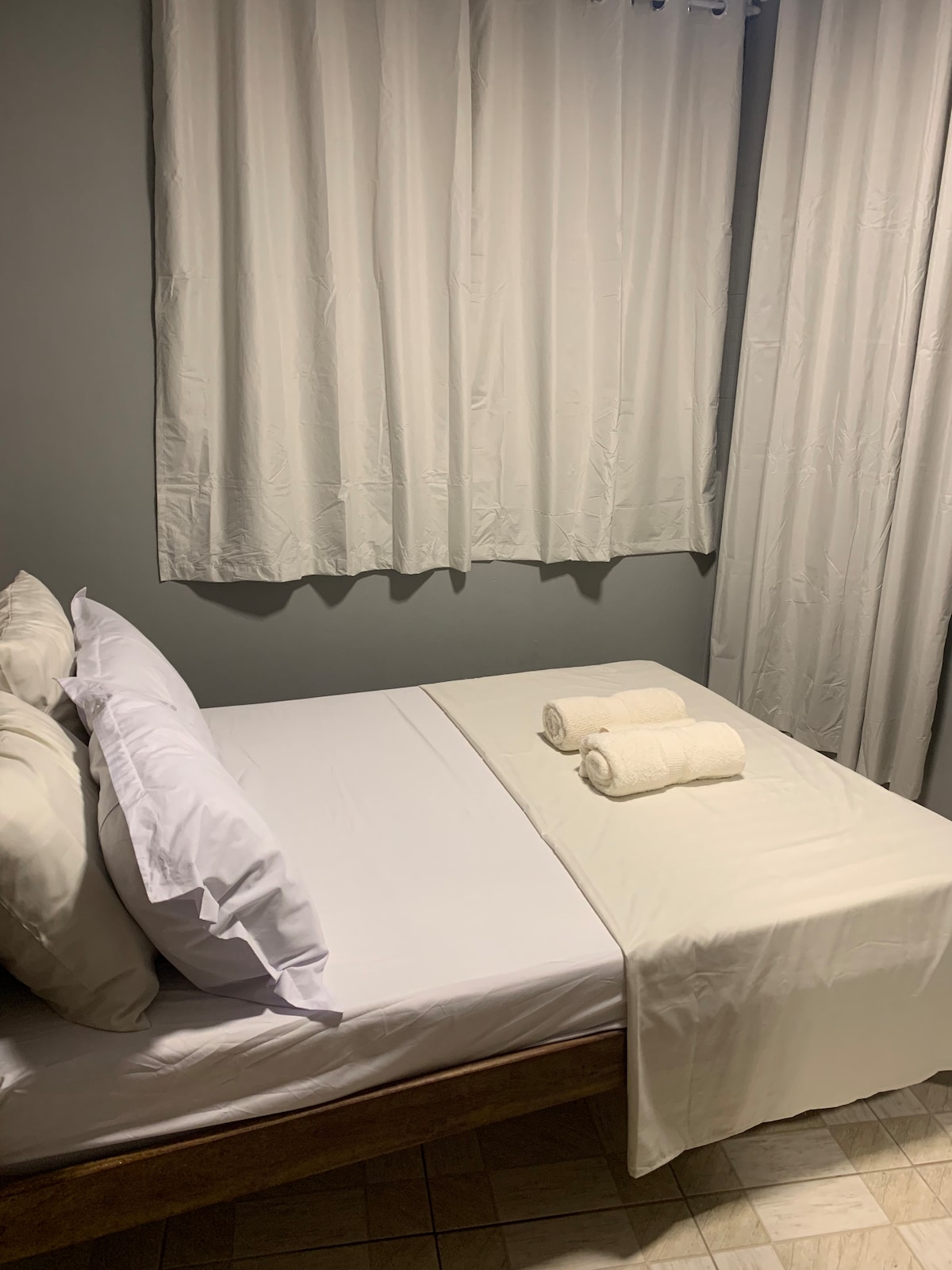 A cozy sleeping area features a neatly made bed with crisp white linens and two rolled towels placed at the foot. Light-colored curtains frame a window, contributing to a bright and airy atmosphere.