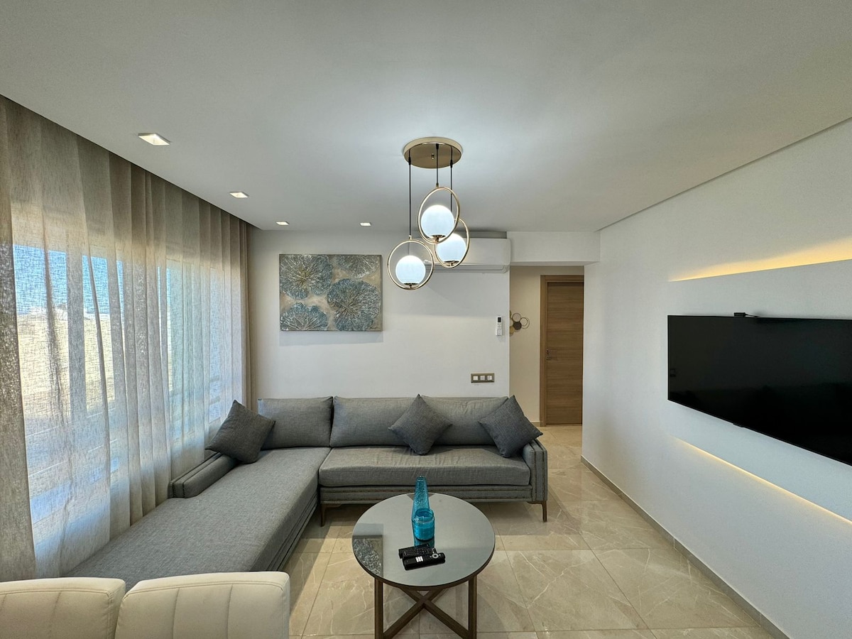 A relaxing living area is presented with a spacious gray sectional sofa, complemented by a glass coffee table. Soft lighting from a modern pendant lamp enhances the ambiance, while sheer curtains allow natural light to filter in from the large windows. A wall-mounted television is also visible.