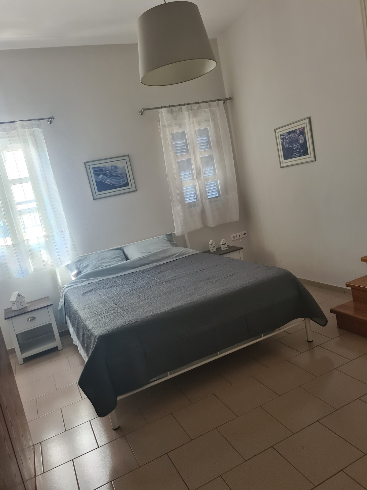 A well-lit bedroom features a spacious bed covered with a soft, grey blanket. Two bedside tables are positioned beside the bed, each with a lamp. Natural light filters through sheer curtains over a window with blue accents, enhancing the calm atmosphere.
