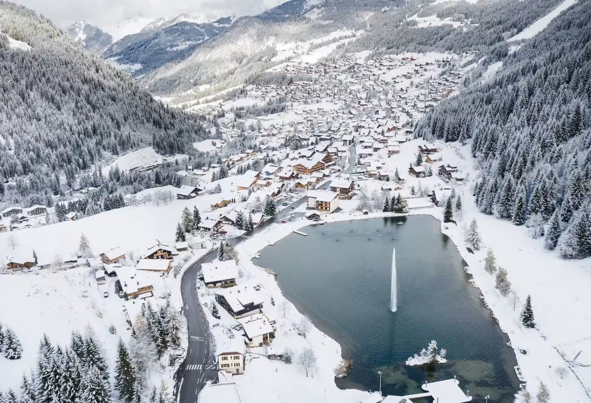 An aerial view captures a snow-covered landscape, showcasing a charming village nestled among mountains. A serene lake reflects the surroundings, with a fountain centered in the water. Snow-draped rooftops extend across the scene, highlighting the peaceful winter ambiance.