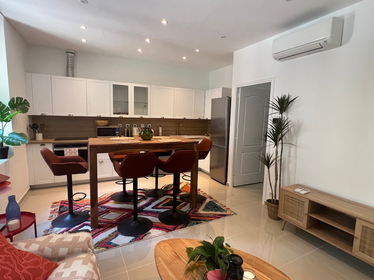 A modern kitchen space features white cabinetry and a wooden countertop. Barstools in red provide seating around a tall table. Appliances include a refrigerator and a stove, while plants add a touch of greenery. Bright lighting enhances the clean and open layout.