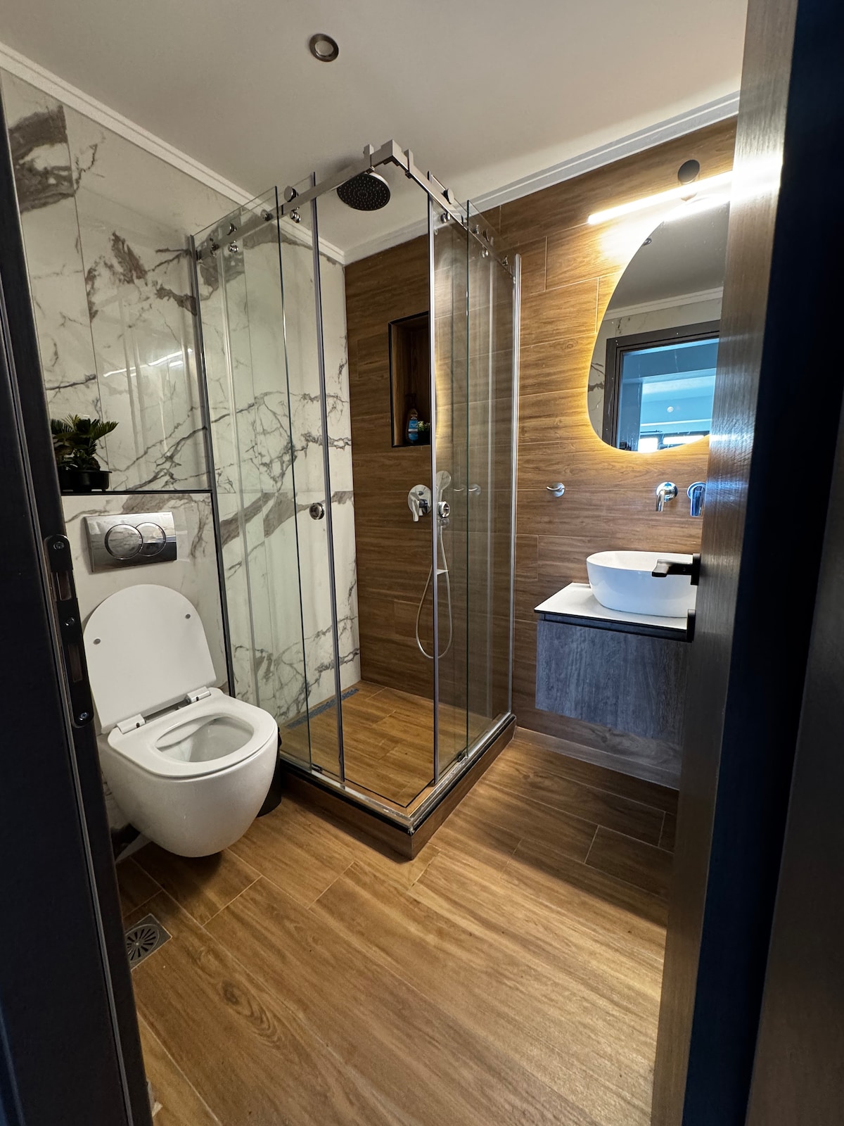 A modern bathroom features a glass-enclosed shower, a stylish circular mirror, and a sleek wall-mounted sink. Natural wood tones are highlighted by well-placed lighting, and marble accents provide a refined touch. A toilet is positioned discretely, completing the contemporary design.