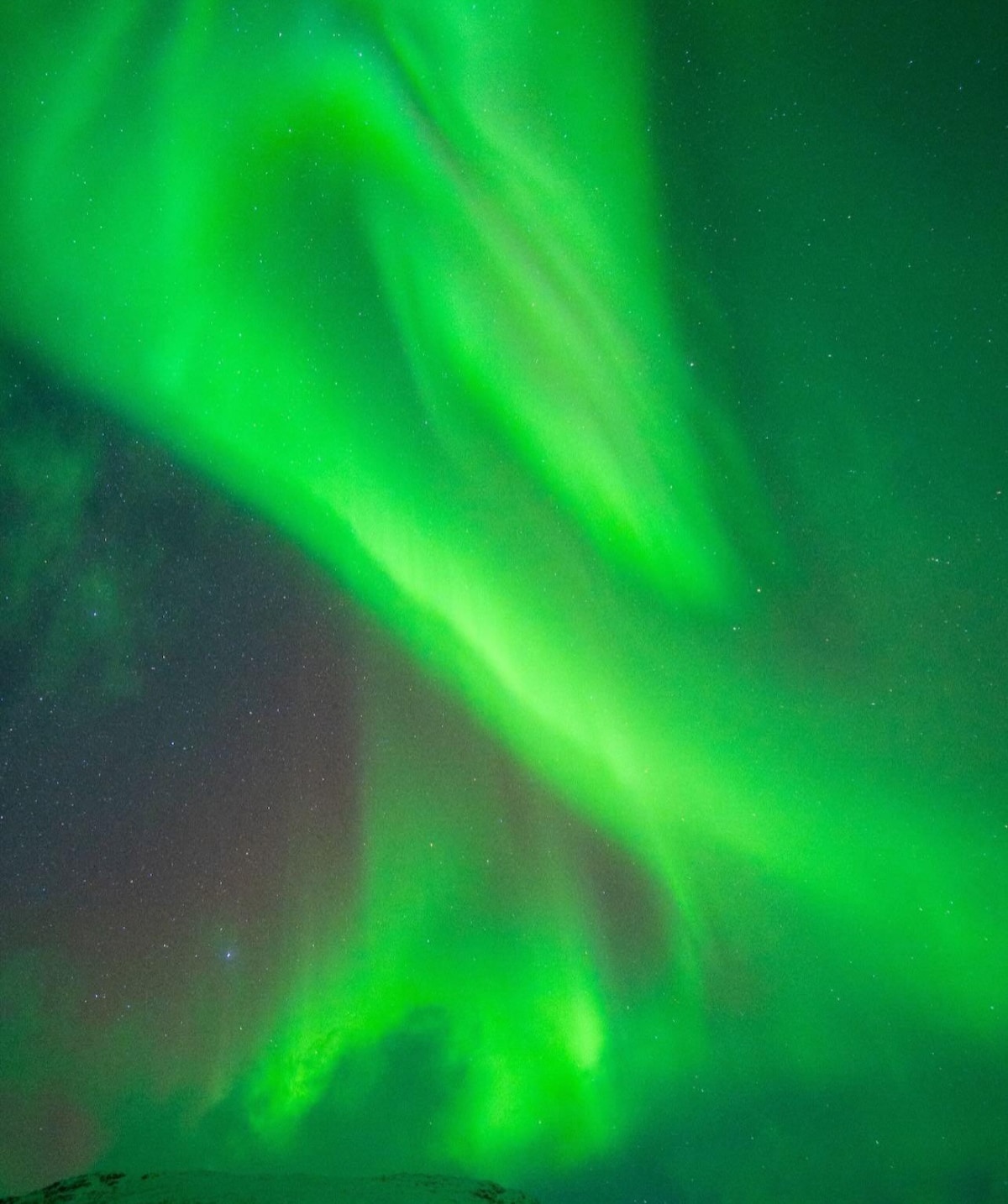 A vibrant display of the northern lights spreads across the night sky, featuring swirling shades of green intertwined with hints of purple and blue. Stars are faintly visible in the dark backdrop, creating a serene celestial scene.
