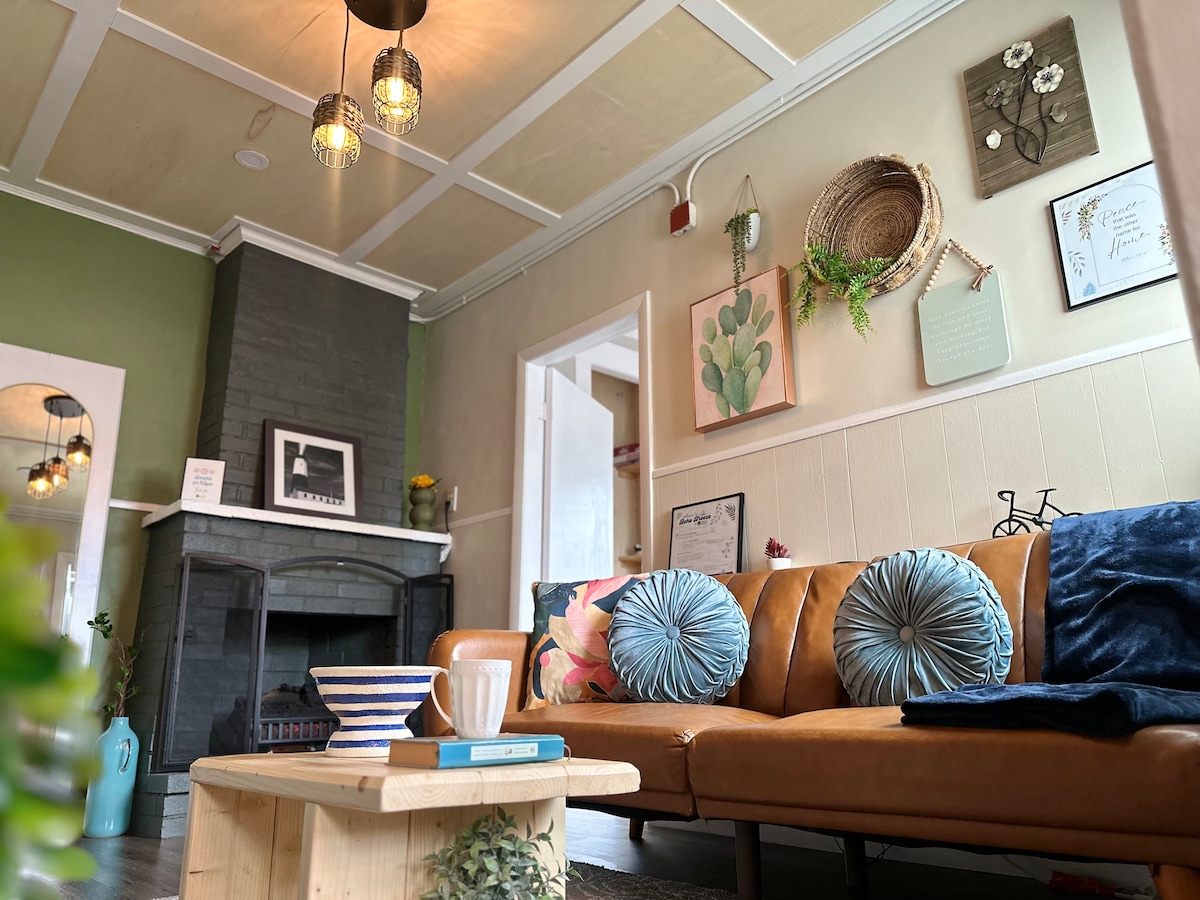 A cozy living area features a brown leather sofa adorned with blue circular cushions. A light wooden coffee table sits in front, complemented by a white mug and decorative bowl. Natural light illuminates the space, highlighting the green painted walls and stylish wall art.