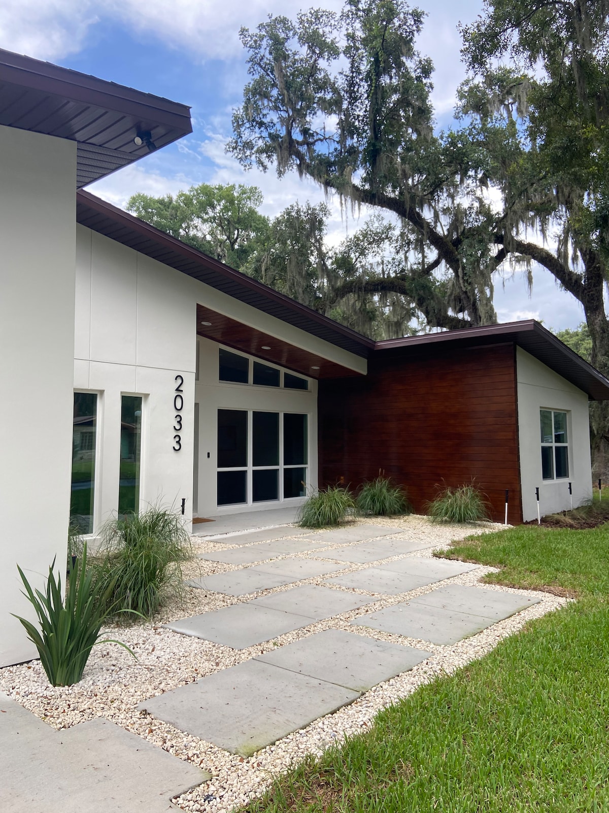 The entrance of a modern home is depicted, featuring a sleek design with a combination of textured white and wood panels. A stone pathway leads through manicured landscaping, bordered by lush green grass and ornamental plants, under a canopy of large oak trees draped with Spanish moss.