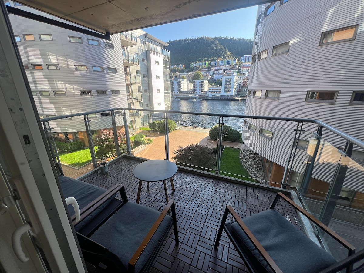 A private balcony offers a serene view of the water and surrounding buildings. Two chairs and a small round table are positioned on the wooden deck, inviting relaxation. Lush greenery is visible below, accompanied by the gentle reflections of nearby structures on the calm water.
